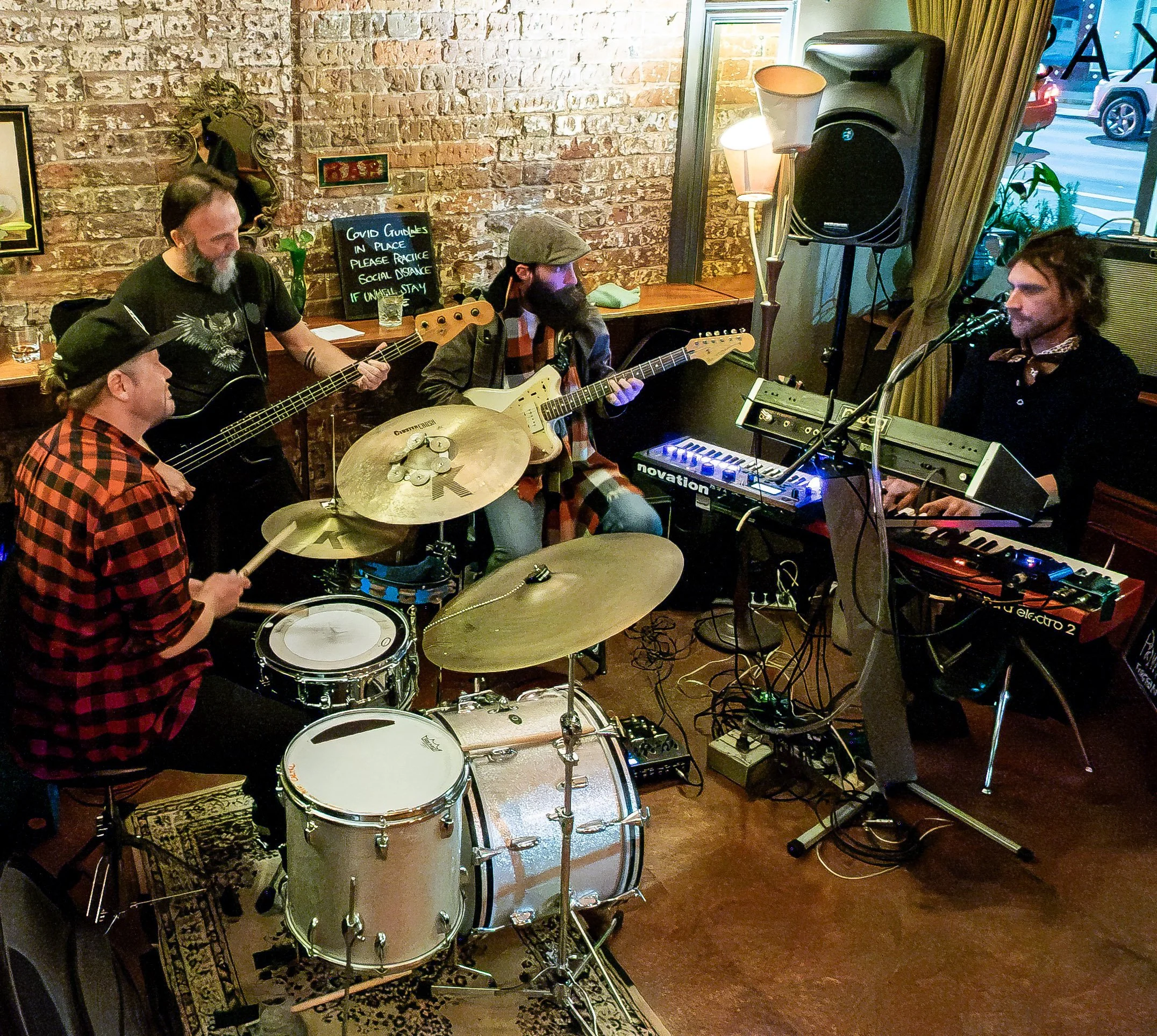 A group of four musicians performing indoors with brick walls in the background. One person is playing drums, others are playing guitars, and a woman is singing or playing keyboard. There is a sign on the wall about social distancing due to COVID-19,