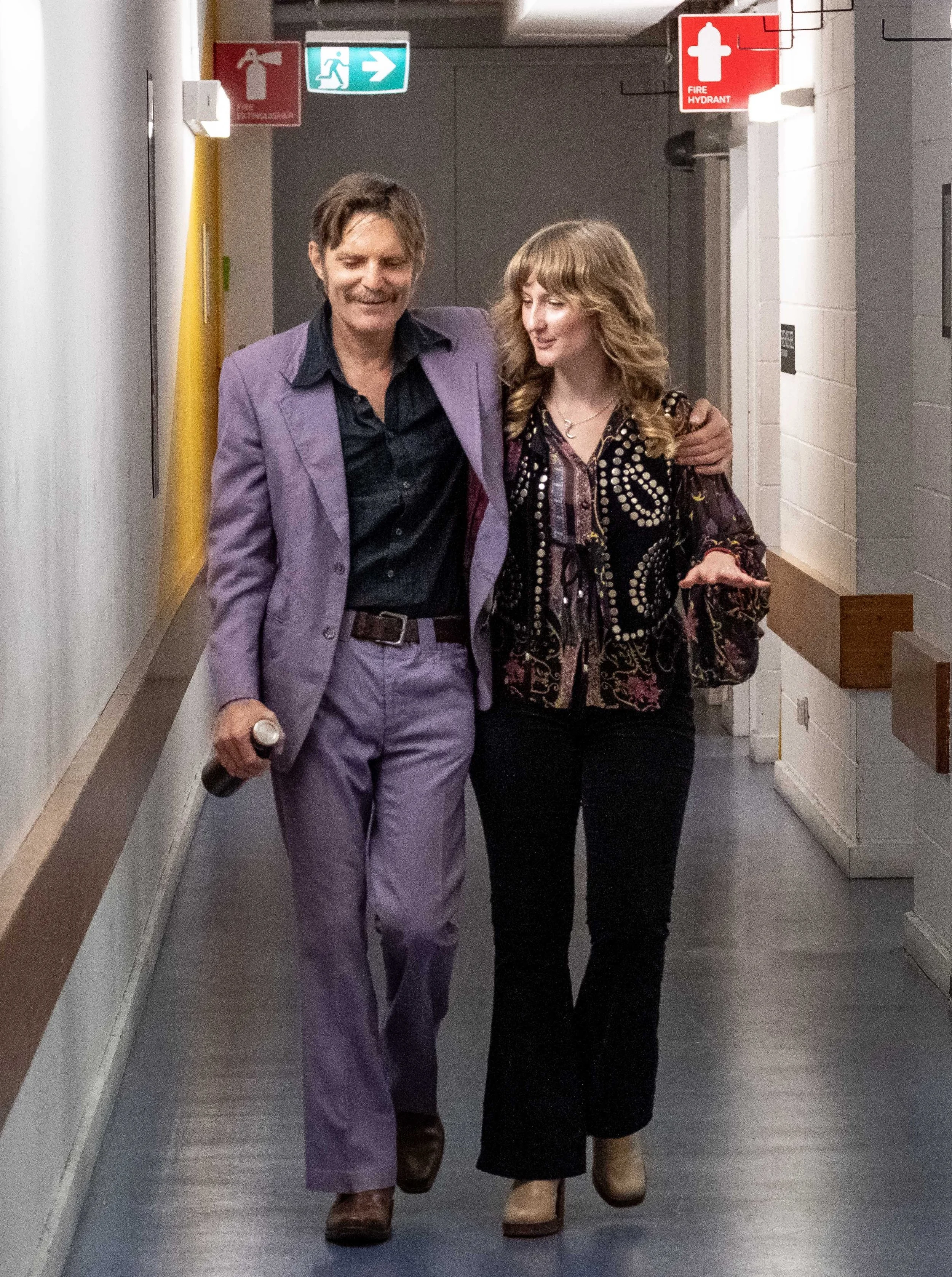 Danny and Dana walking together in a hallway, with emergency exit signs and fire safety signs above, and a beige wall on the left, and a gray wall on the right.