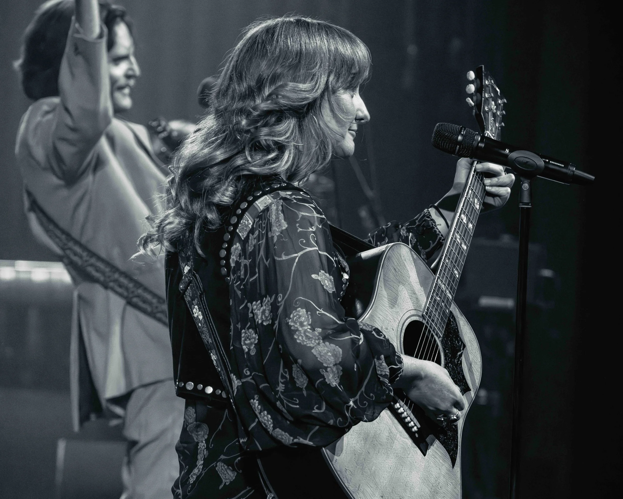 A woman playing an acoustic guitar and singing into a microphone during a performance, with a smiling person in the background.