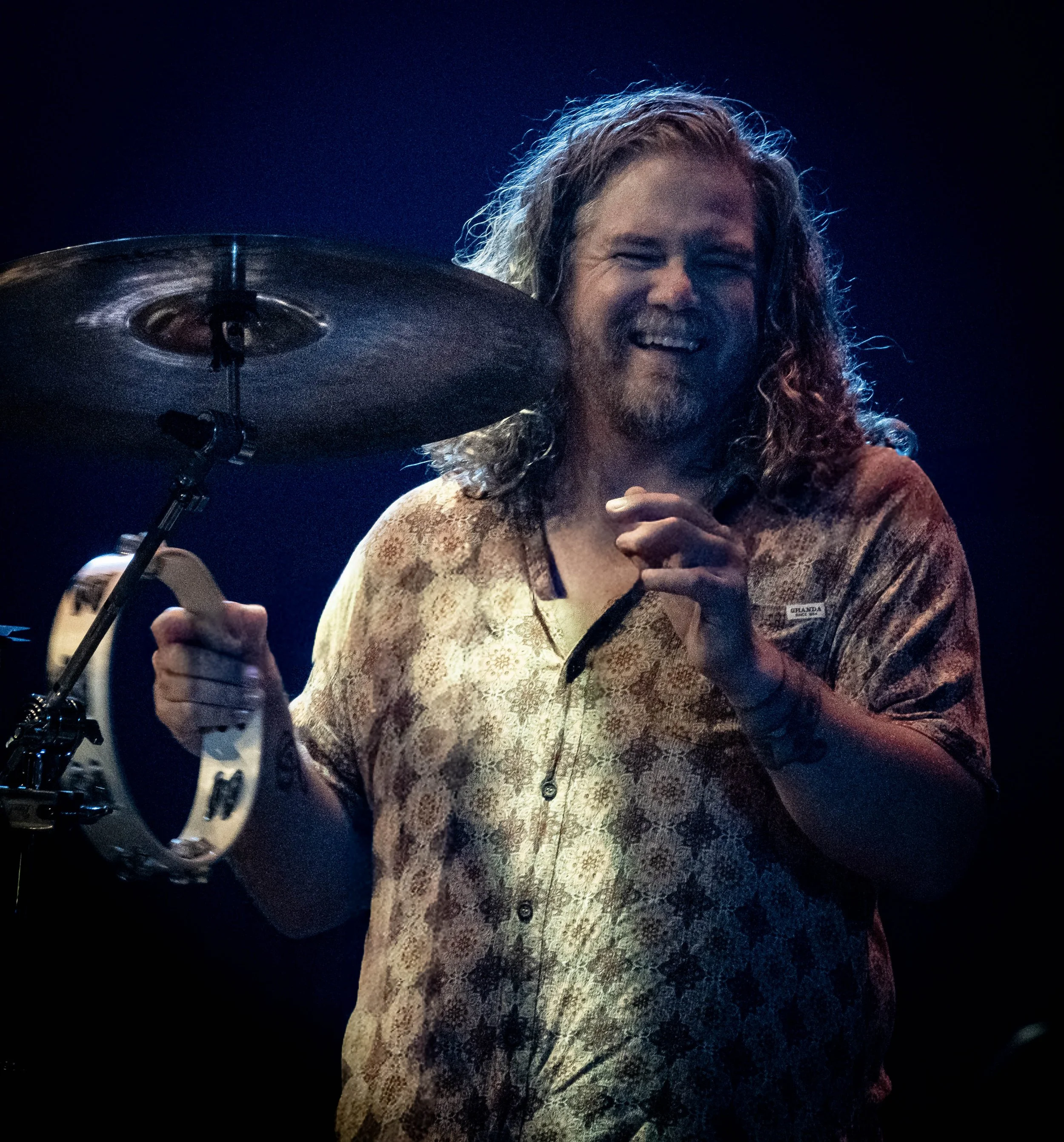 A man with long hair and a beard smiling while playing drums on stage. He is wearing a patterned shirt.