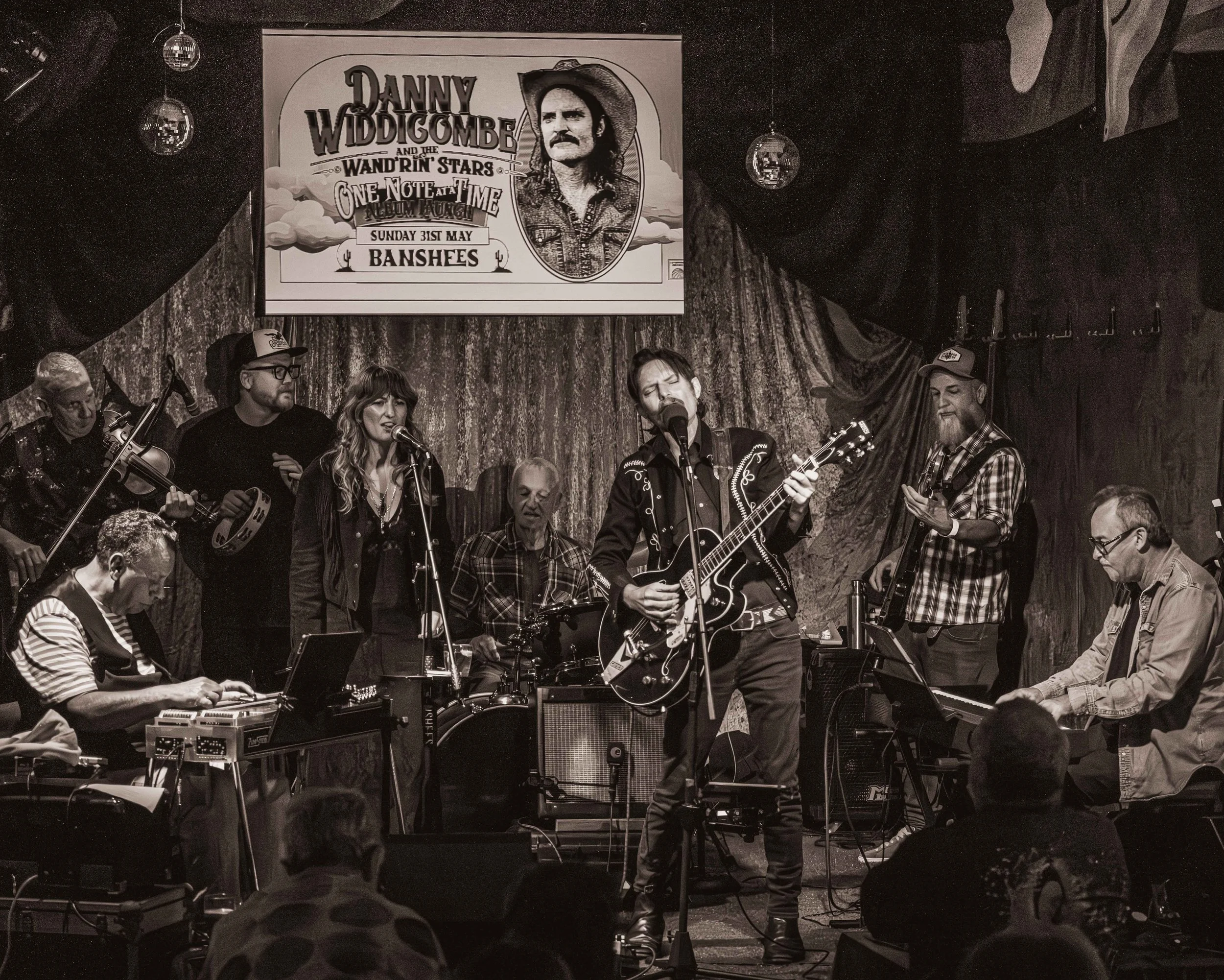 A band performs live on stage, featuring a lead singer with a guitar, a woman singing, and several other musicians playing instruments. A sign above the stage advertises a concert by Danny Widdicombe and the Wandrin' Stars, with a theme titled 'One Note at a Time Album Launch' scheduled for Sunday, May 31st, at Banshees.