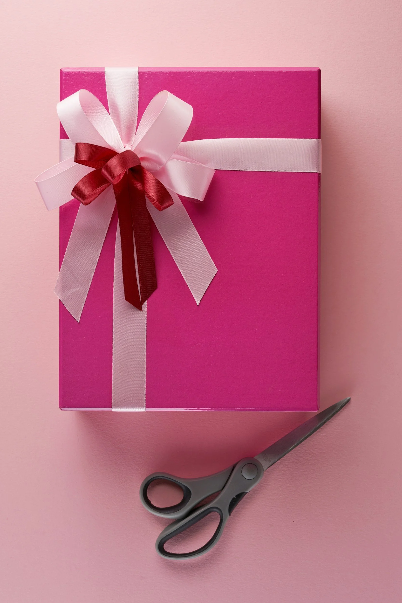 Pink gift box with a large pink and white ribbon bow on top, placed on a pink background. Scissors beside the box.
