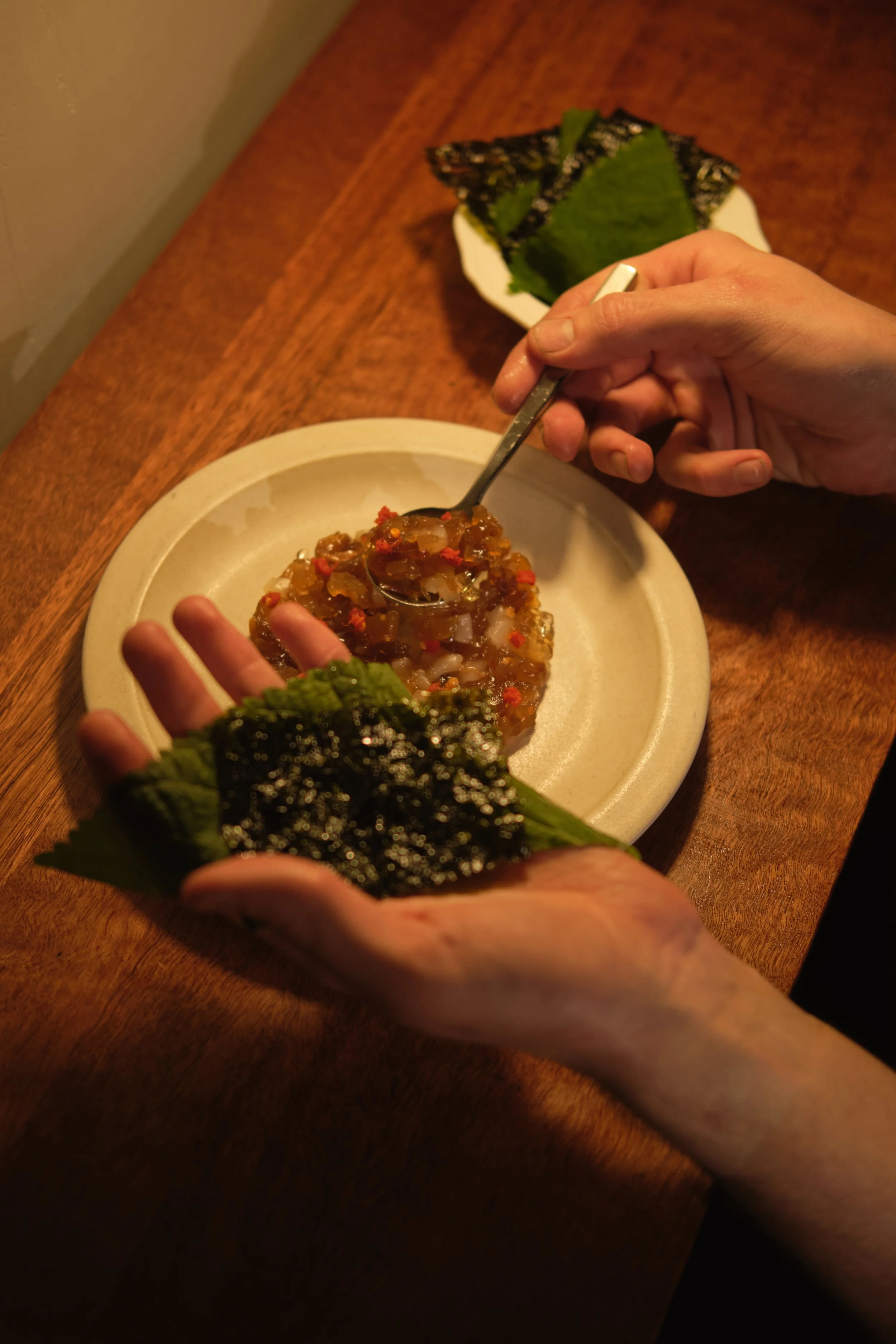 A person holding a piece of nori seaweed over a plate of sashimi with another plate of sashimi in the background. hospitality, food photography, commercial and social media work available now, creative food photographer