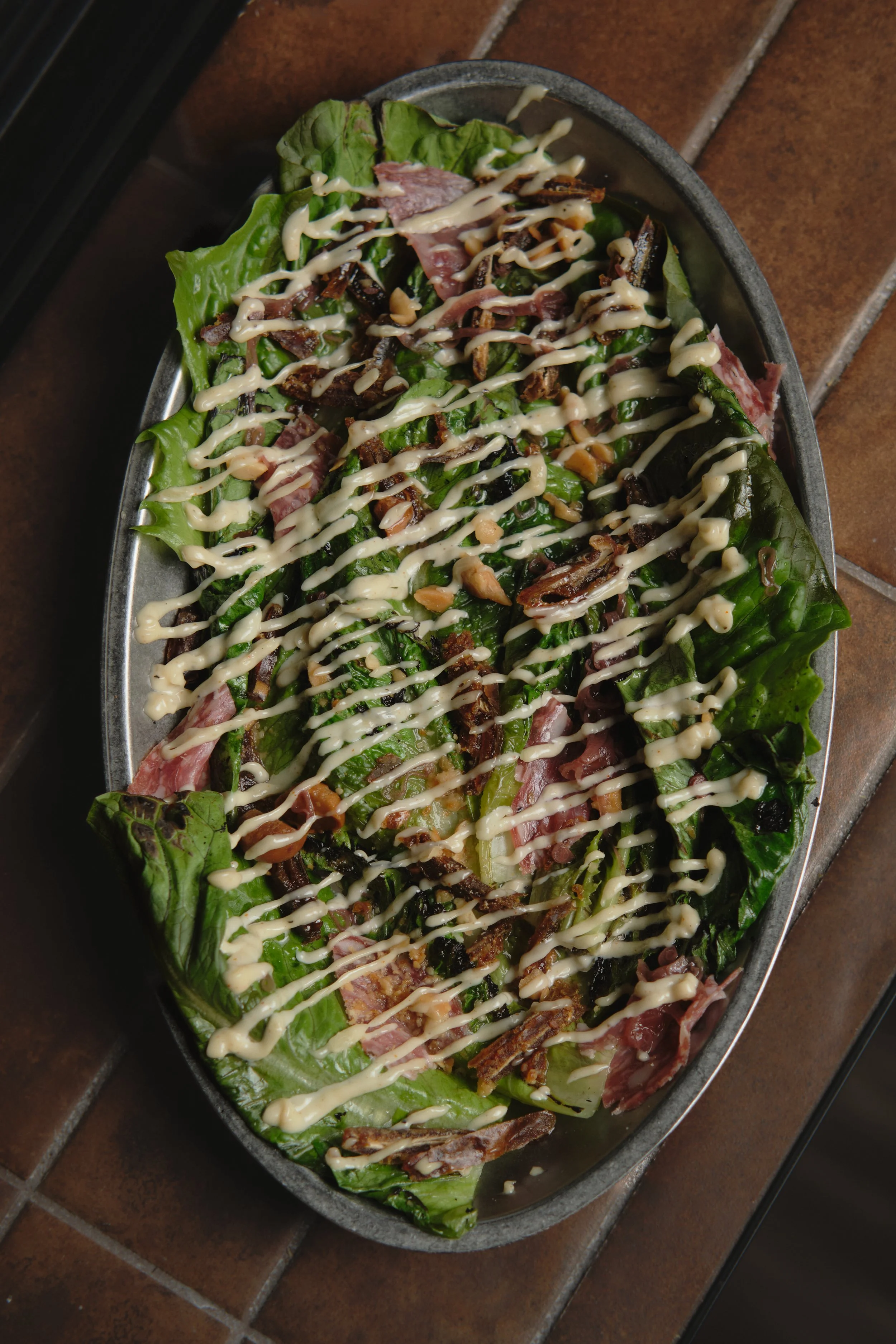 A salad with lettuce, slices of meat, crispy bacon, and chopped nuts, drizzled with creamy dressing, served in a metal dish on a brown tiled surface.photography for hospitality venues, lifestyle, product photography, spirits, beverages, cocktails.