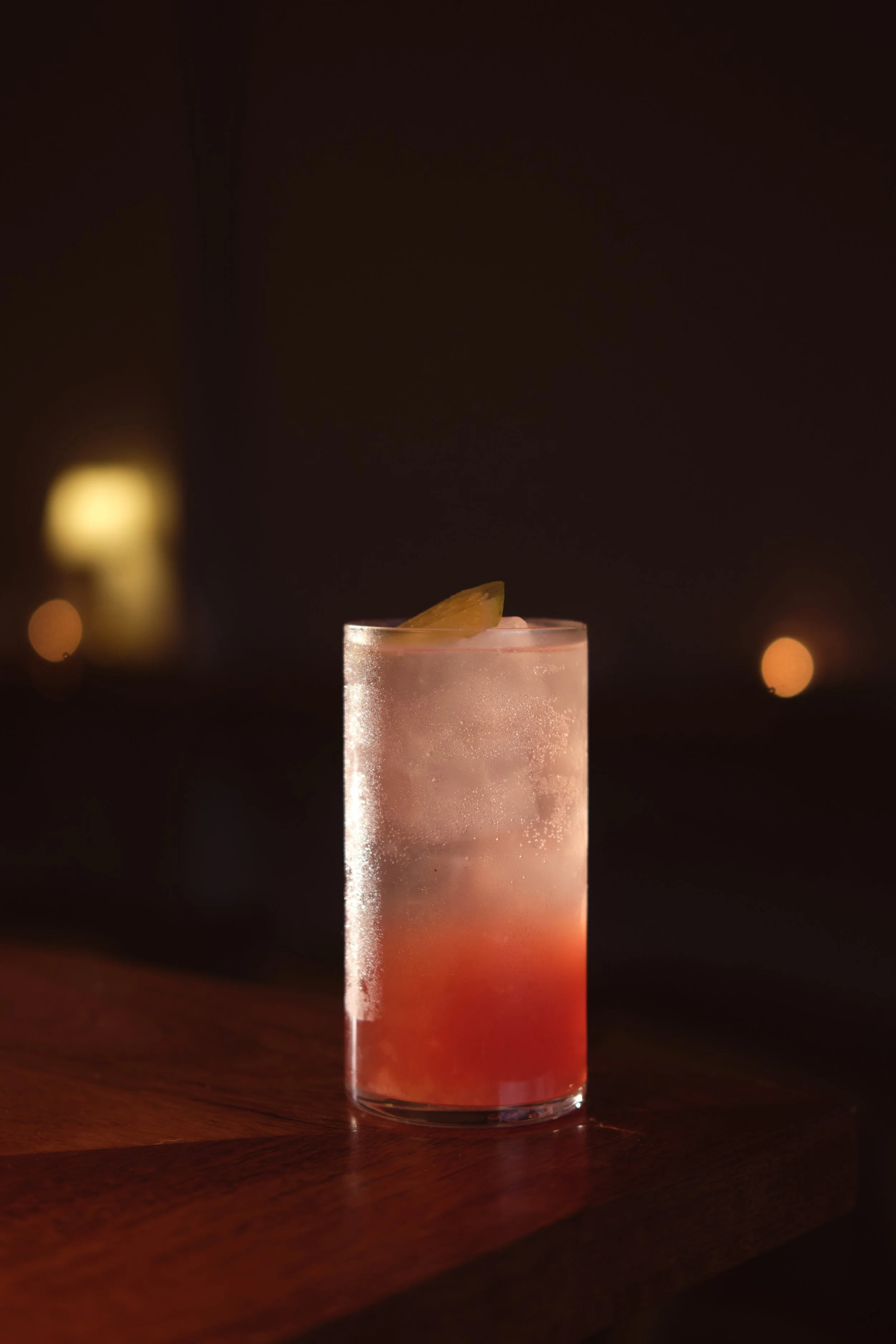 A tall glass cocktail with a gradient of colors from red at the bottom to clear at the top, topped with a slice of lemon, and illuminated in a dark setting with a blurred background of lights. cocktail photography, drinks photography, hospitality.