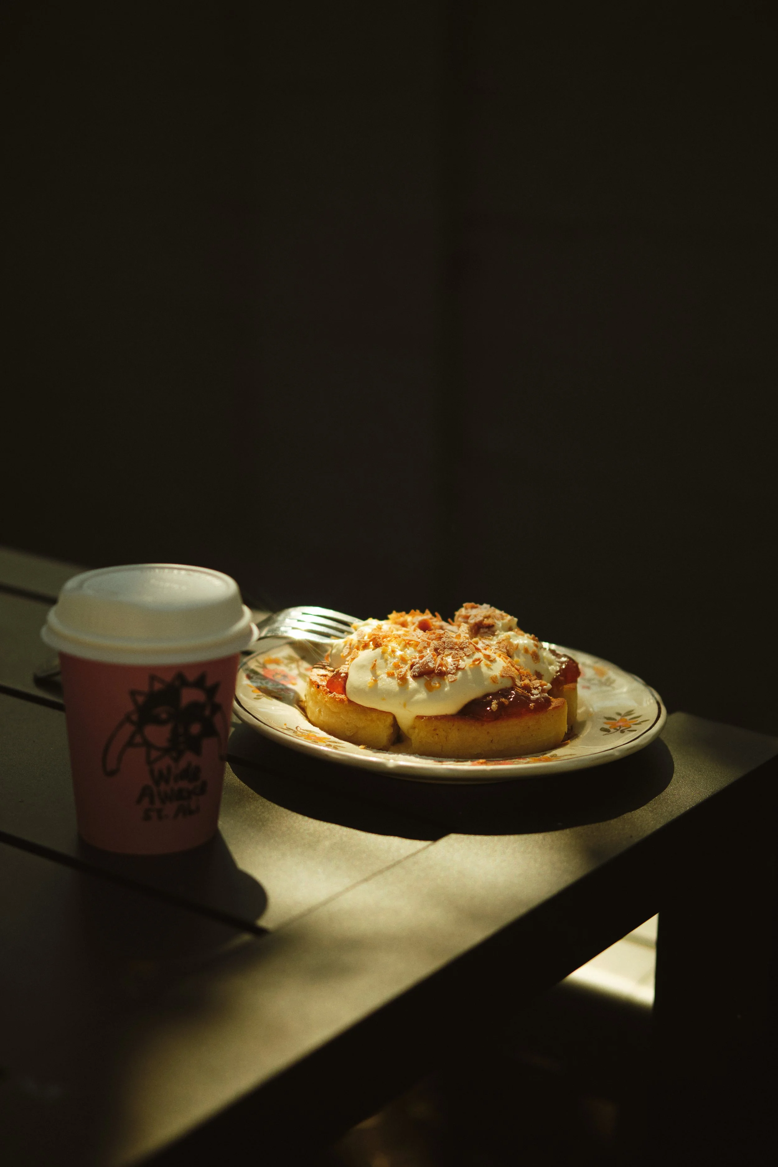 A plate with a slice of cake topped with whipped cream and crumbles, a disposable coffee cup with a hot beverage, and a fork on a table against a dark background. photography for hospitality venues, lifestyle, product photography, cafe
