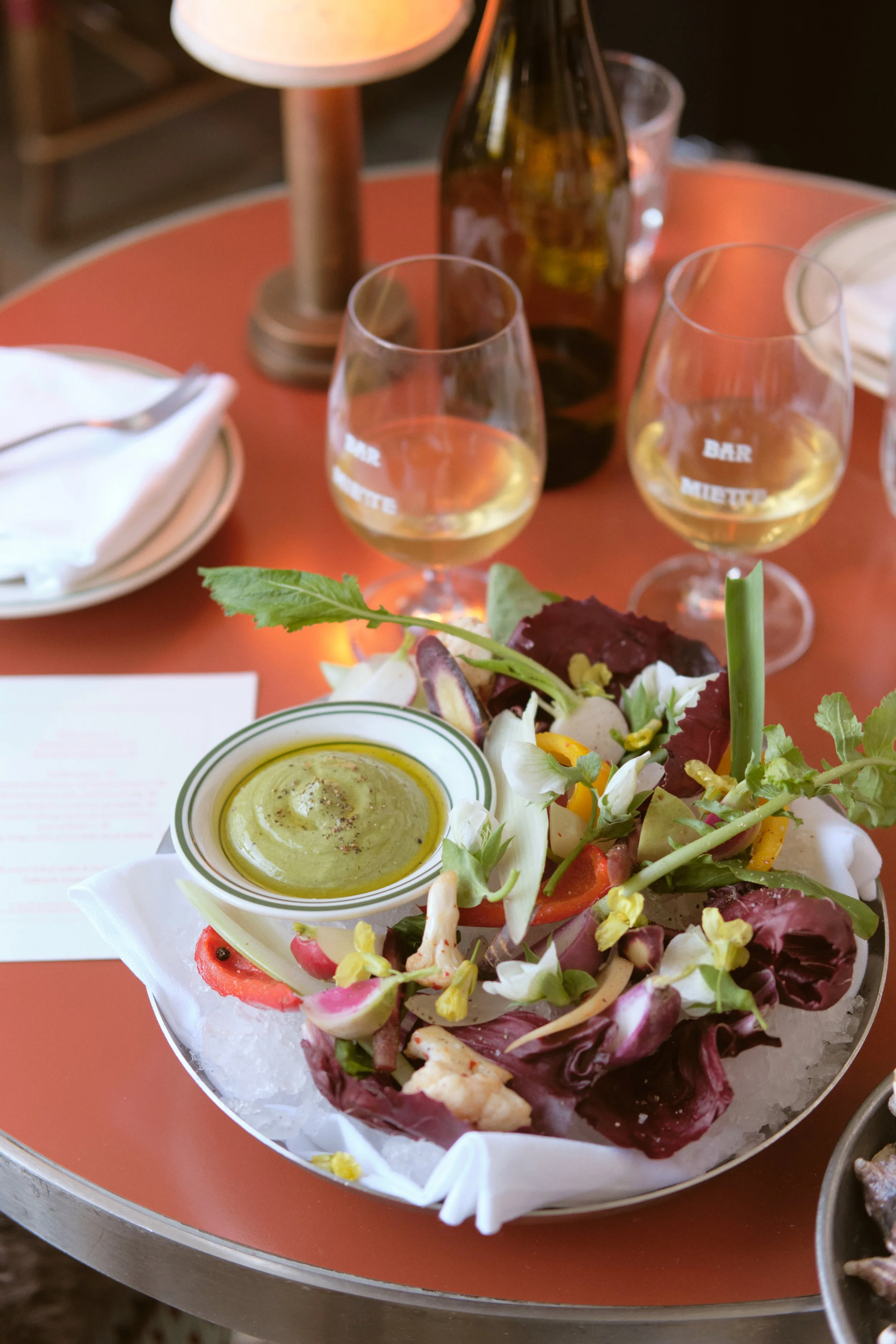 A salad with mixed greens and vegetables topped with a small dish of green dressing, set on a table with two glasses of white wine and a bottle in the background. drinks, food photography for venues, brisbane, australia, travel, hotel photographer.