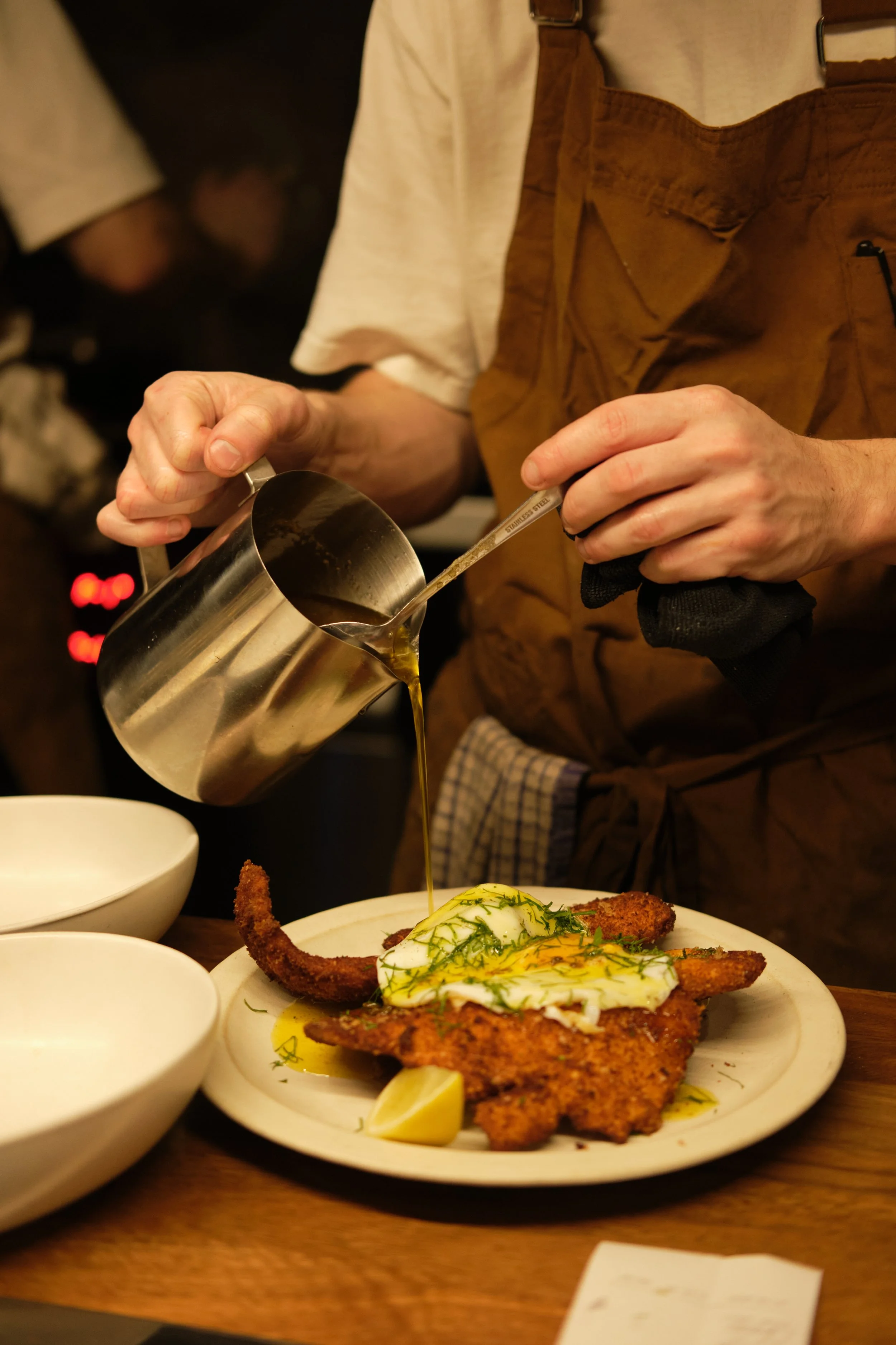 A person pours sauce or oil over a fried fish fillet topped with a fried egg and garnished with dill, served with lemon wedges on a white plate.photography for hospitality venues, lifestyle, product photography, spirits, beverages, cocktails, food.