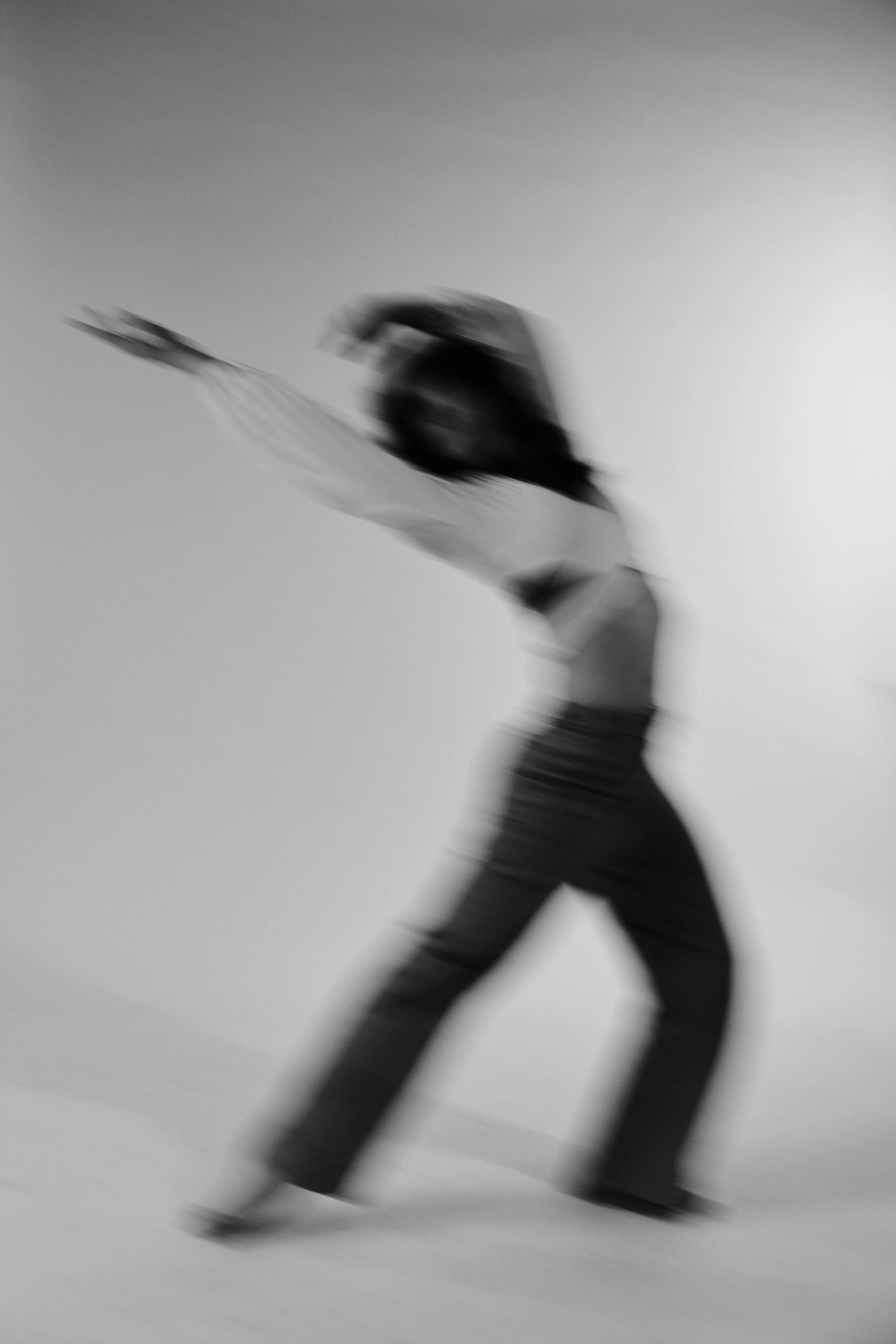 A blurred black and white photo of a person with long hair walking or running against a plain background, with motion blur and no clear details.