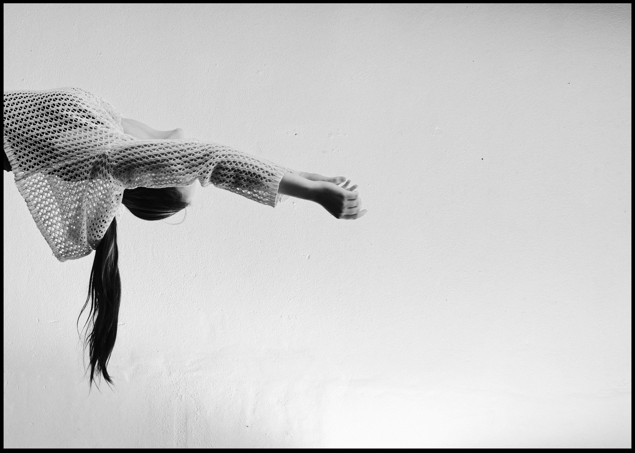 A black and white photo of a girl with long hair in a ponytail, wearing a wide-knit sweater with open stitching, reaching out with her right hand against a plain wall, creative circus arts photography. 