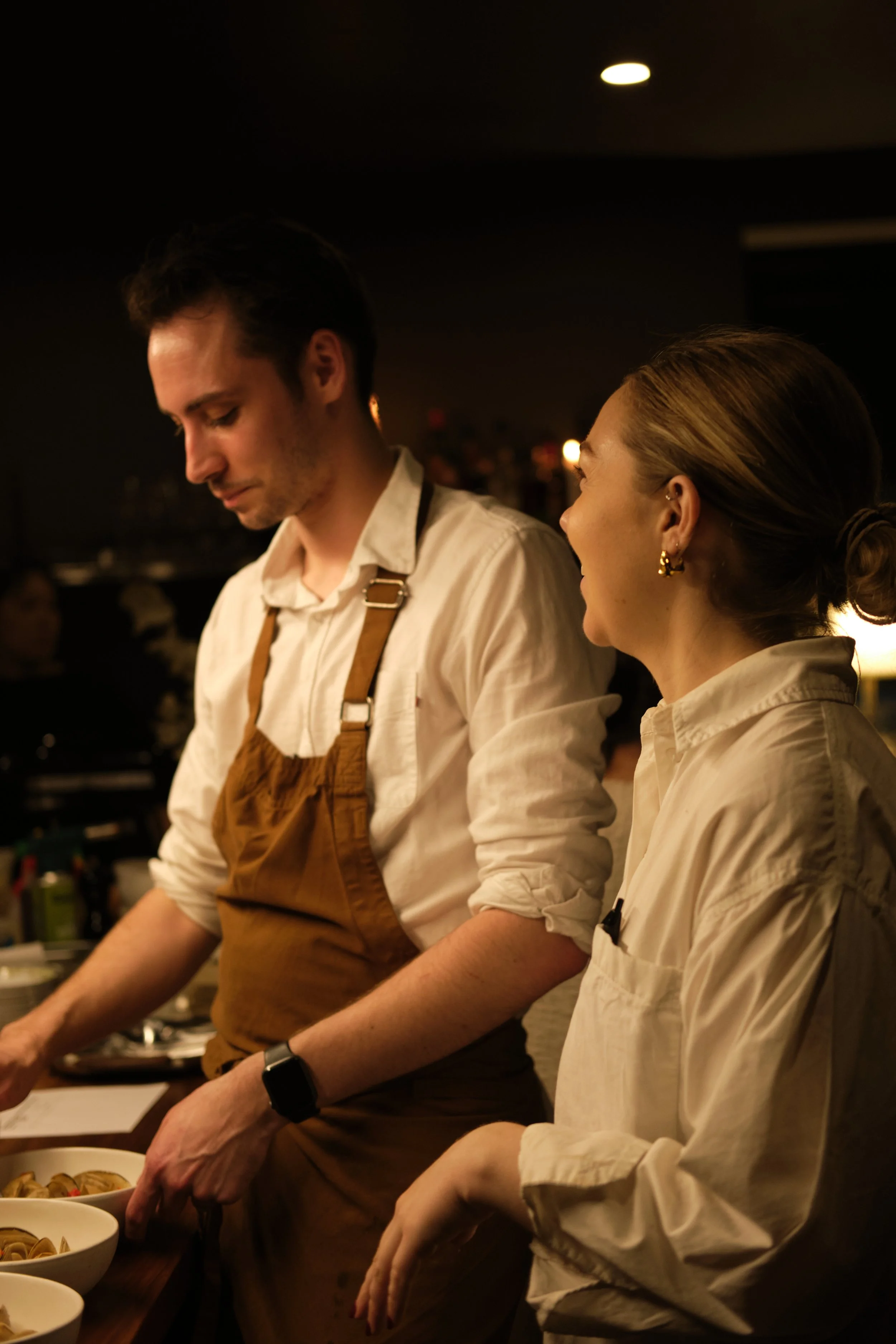 Two people working behind a counter in a dimly lit restaurant kitchen, with bowls of food on the counter, engaged in preparing or serving food. portrait photography, hospitality, service,.