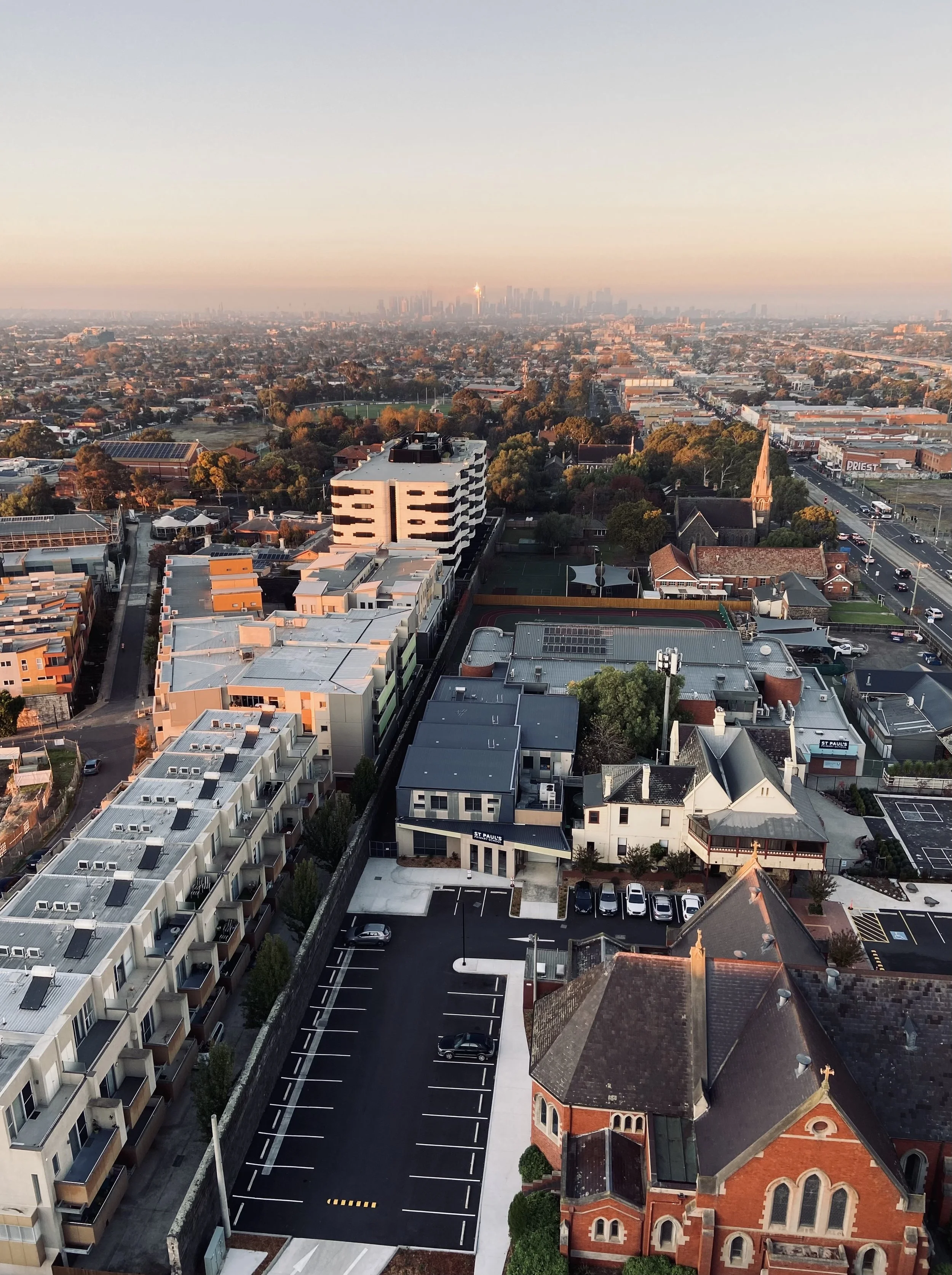 Aerial view of a cityscape during sunset with a church in the foreground, residential and commercial buildings, parking lot, and distant skyscrapers on the horizon.