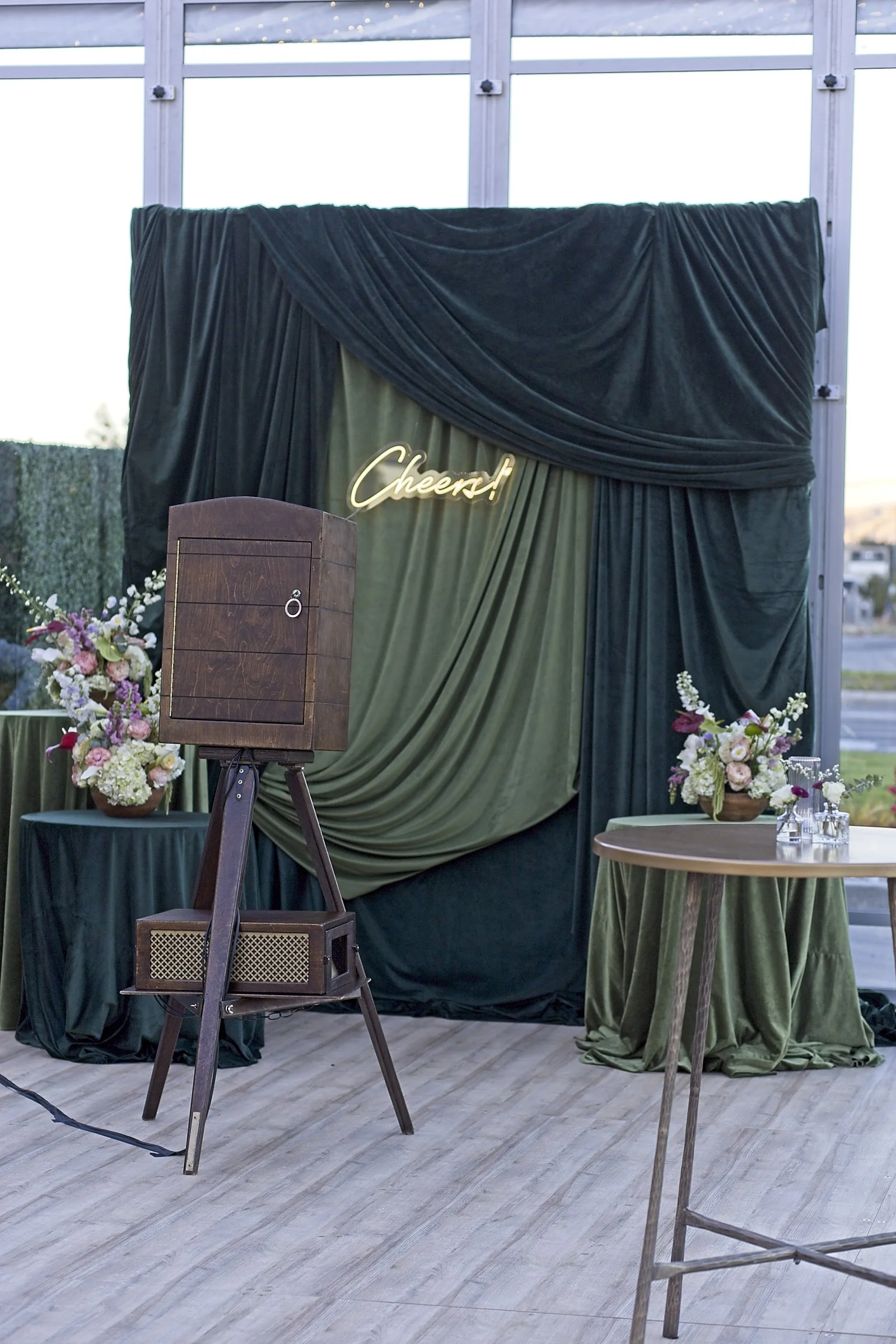 Indoor event setup with dark green draped backdrop, potted flower arrangements, a wooden podium, a vintage wooden speaker, a small table with a green tablecloth, and a table with glasses, all under large windows and a metal structure.