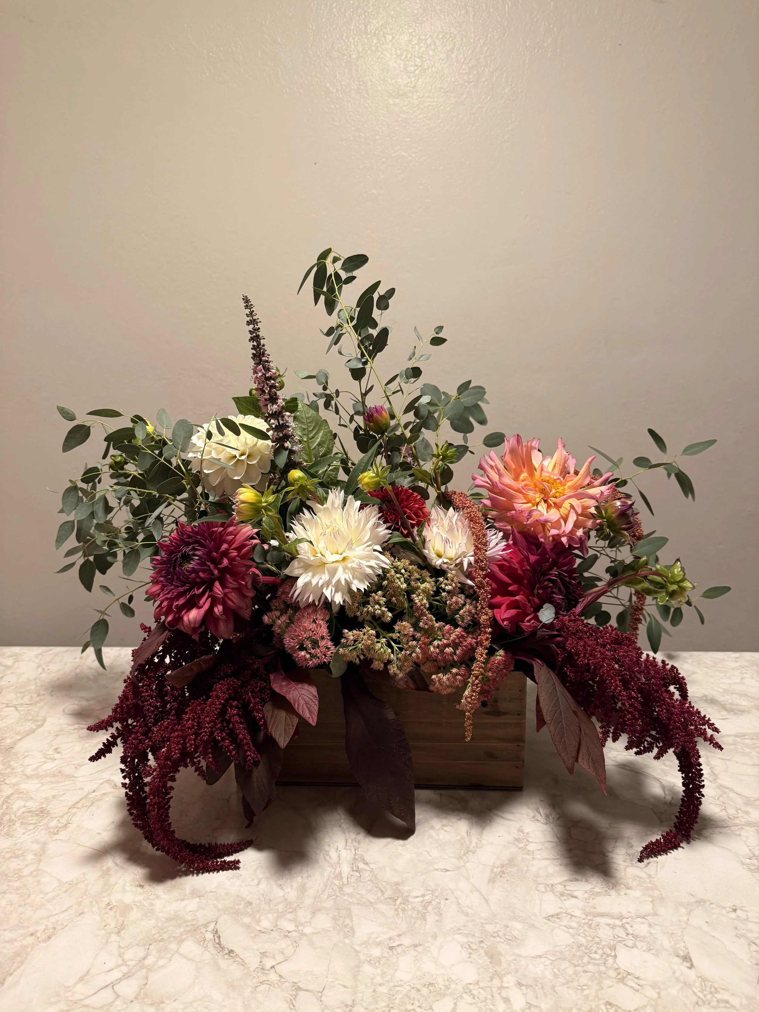 A floral arrangement with various colorful flowers, greenery, and foliage arranged in a wooden box on a marble surface against a beige wall.