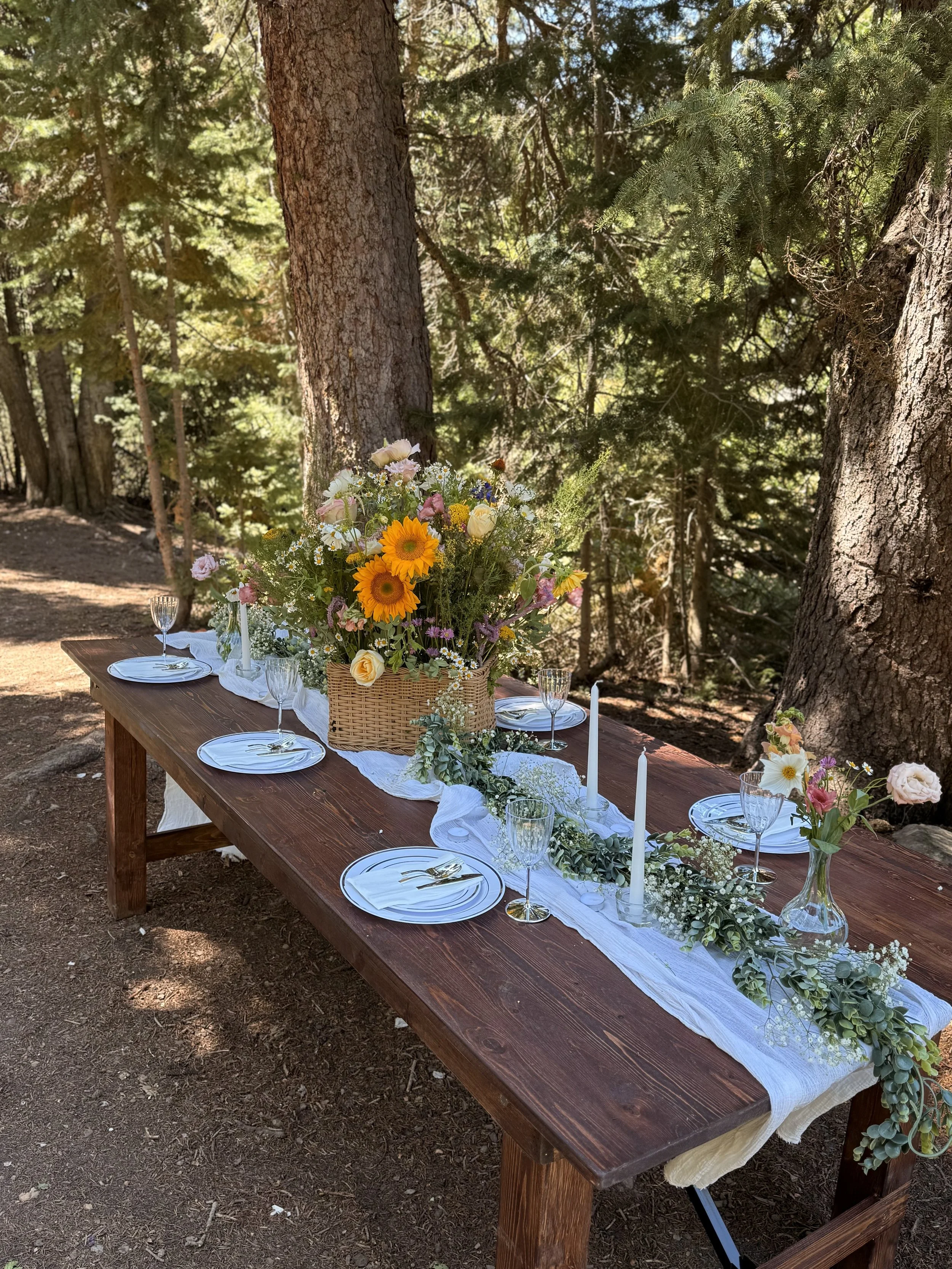 A wooden outdoor table set for a meal, decorated with a white table runner, floral centerpiece with sunflowers and assorted flowers, white candles, plates, glasses, and cutlery, situated in a forest setting with tall trees and sunlight filtering through the leaves.