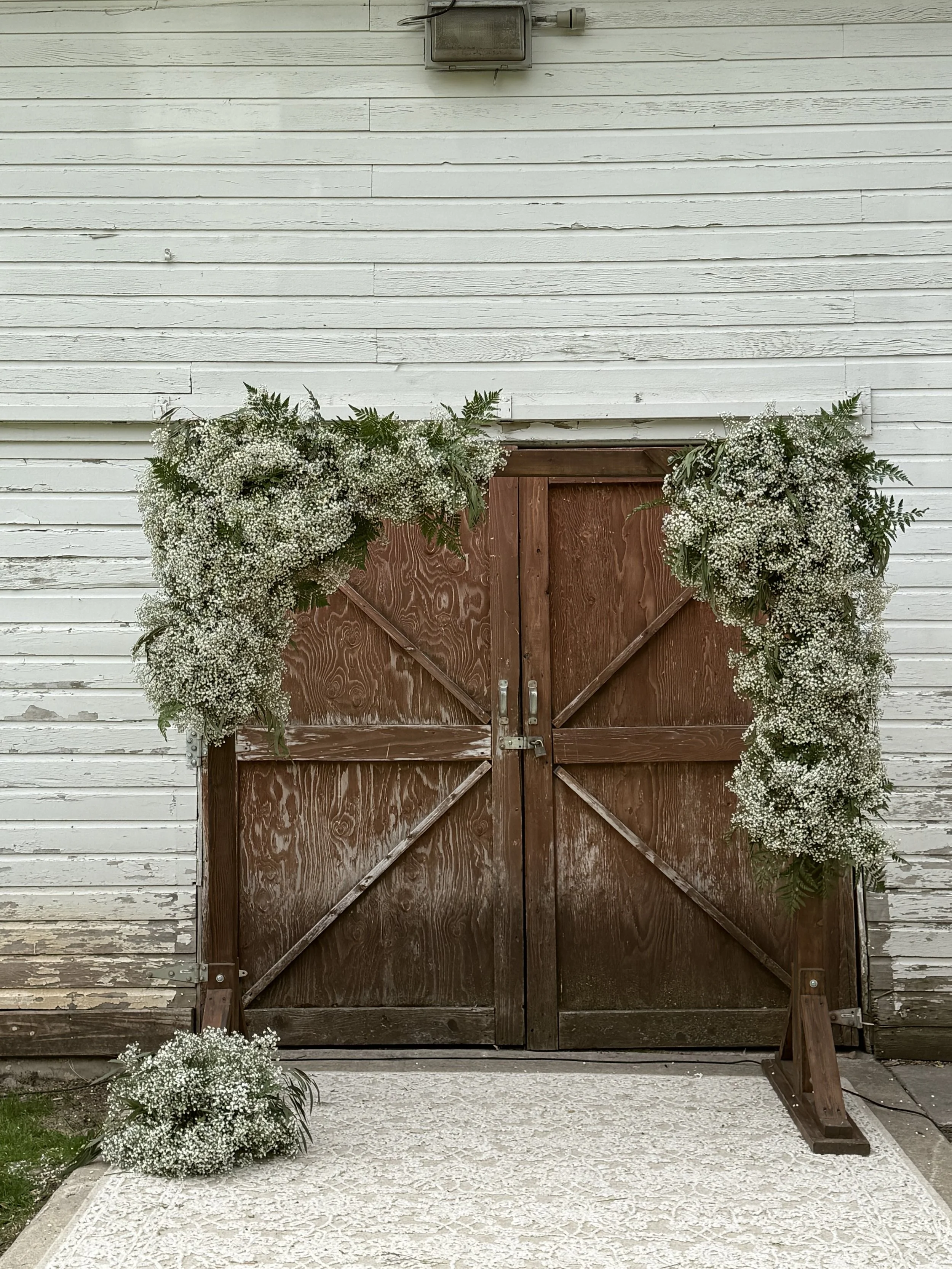 A wooden barn door decorated with white flowers and greenery, with a white wooden wall in the background and a small flower arrangement on the ground in front.