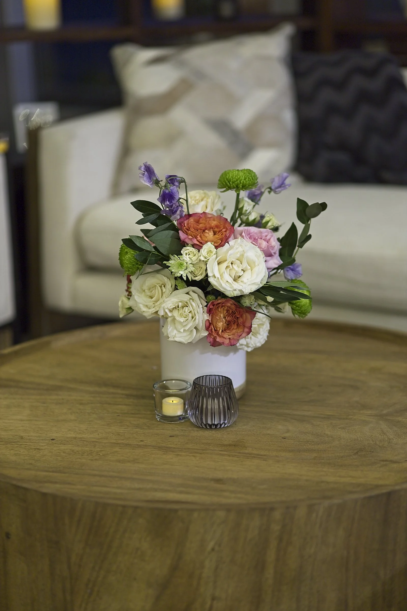 A round wooden table with a white vase of mixed flowers including white, pink, orange, purple, and green blooms, two small candles in glass holders, and a purple ribbed glass tumbler in a cozy indoor setting with pillows and a shelf in the background.