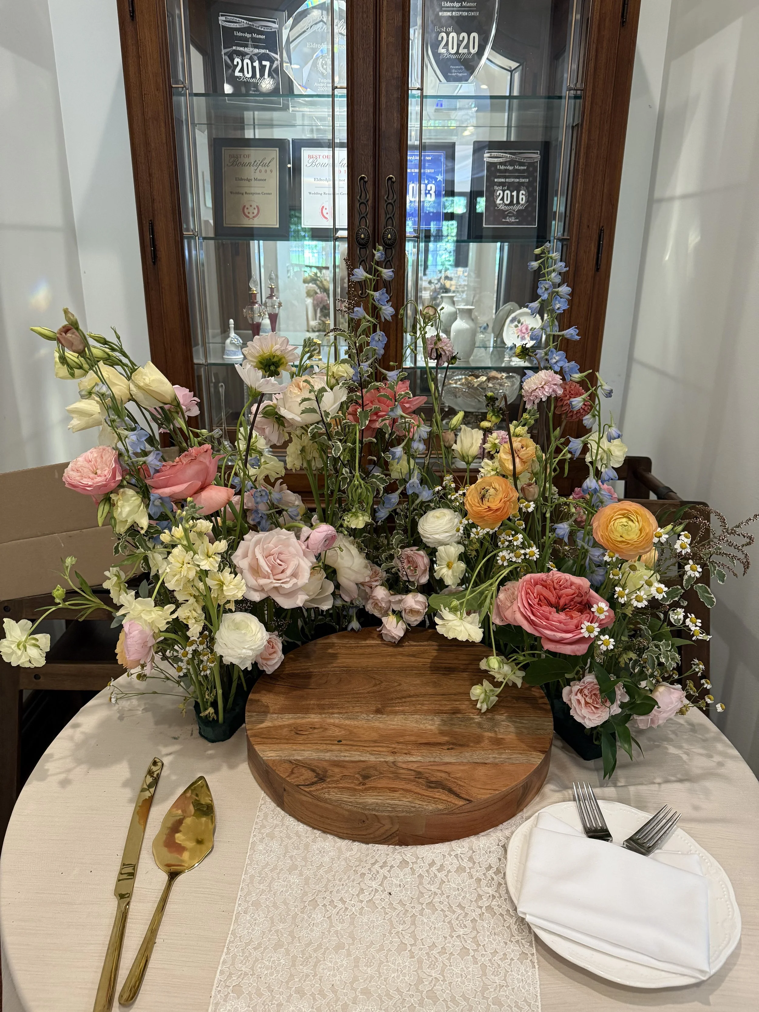 A table with a large floral centerpiece, a wooden serve platter, a white plate with a folded napkin and forks, and gold-colored serving utensils, set for a meal.