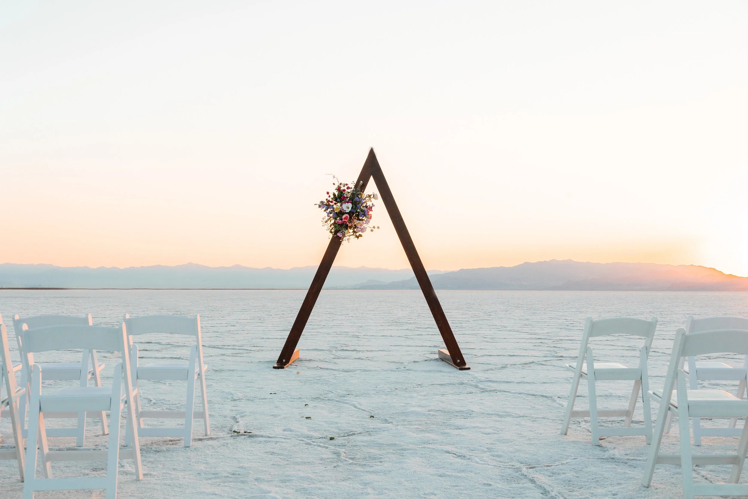 A wooden wedding arch decorated with colorful flowers is set up on a white sandy beach at sunset, with white chairs arranged for an outdoor ceremony overlooking a calm body of water and distant mountains.