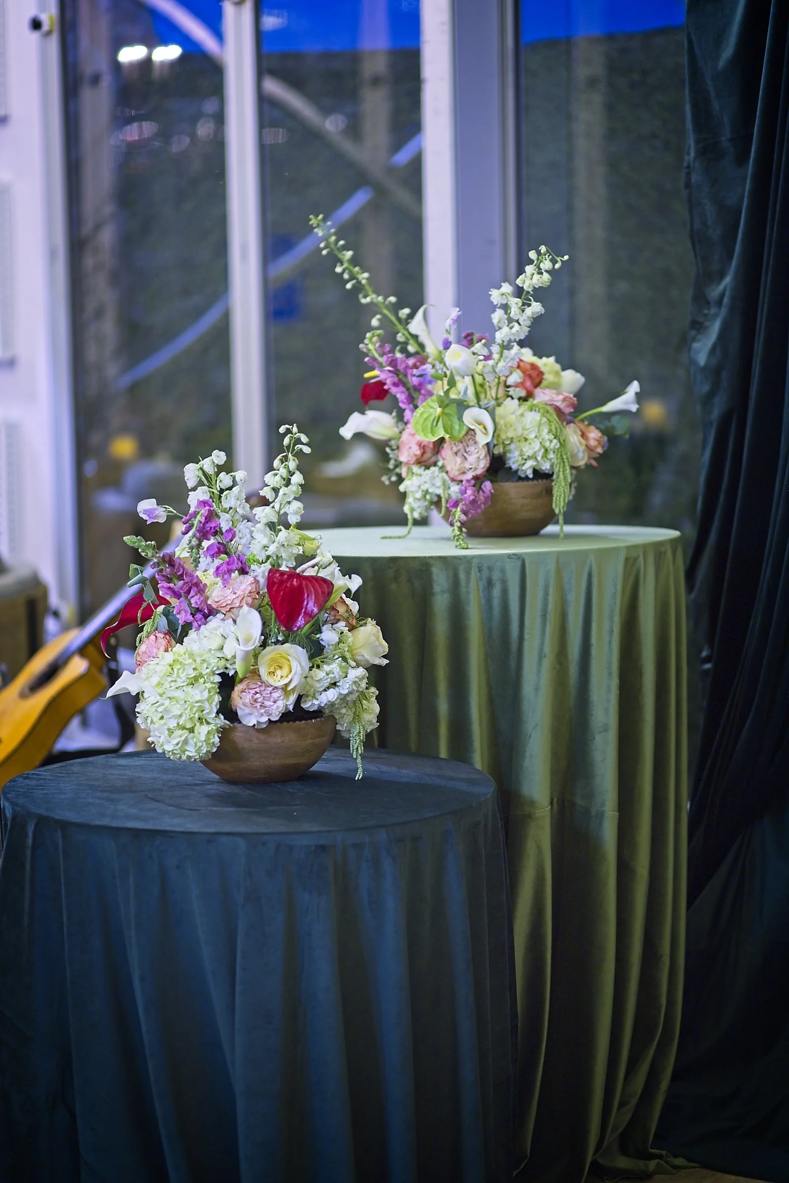 A floral arrangement in a wooden bowl on two tables covered with dark green and black tablecloths, set inside a venue with a window and curtains.