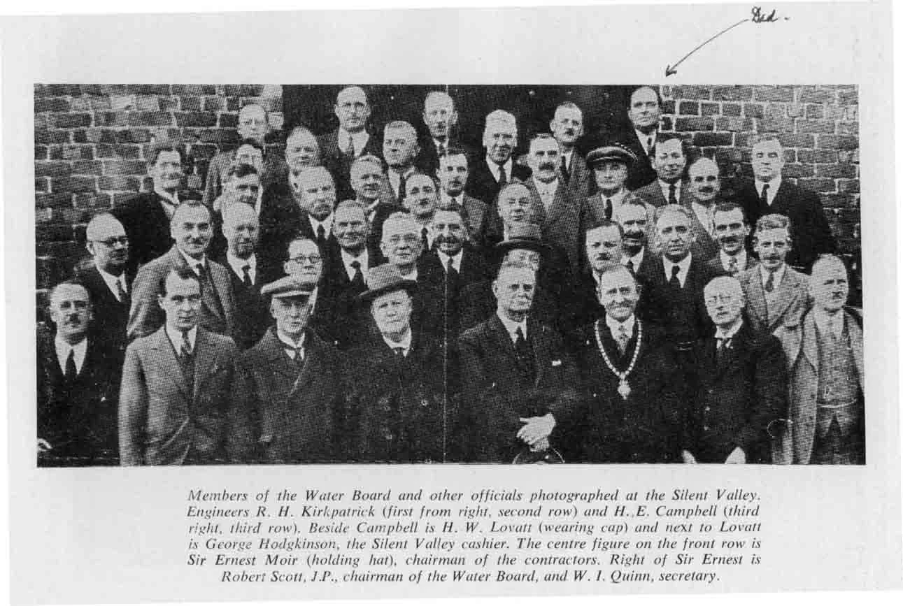 Black and white photograph of a group of men and women standing in front of a brick wall, dressed in early 20th-century clothing. Some are wearing hats, and one woman is wearing a necklace. There is a handwritten note and arrow pointing to the top right corner of the photo. The caption underneath explains it shows members of the Water Board and other officials at the Silent Valley, with details about specific individuals in the front row.