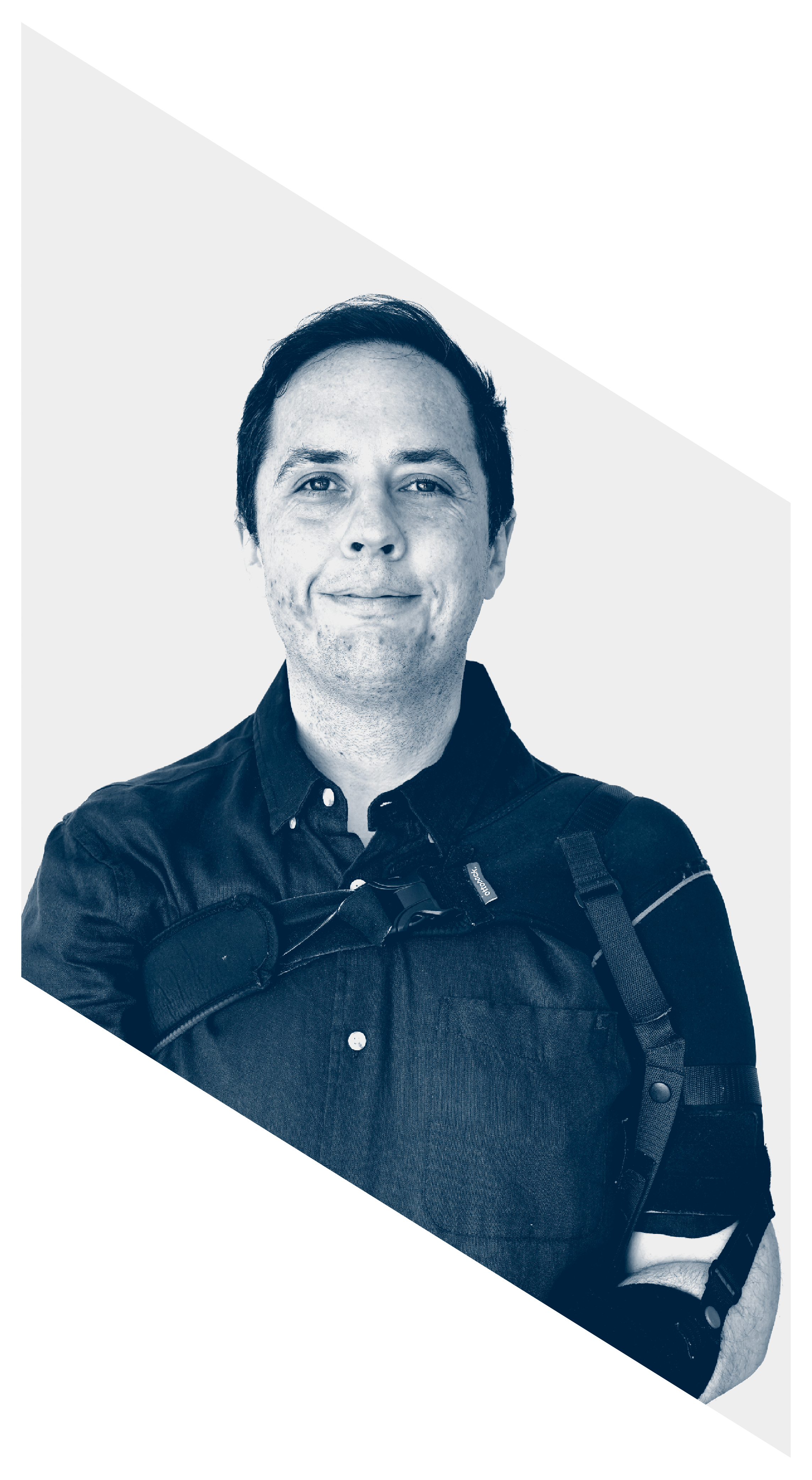 A man with short dark hair, wearing a black button-up shirt, smiling slightly, carrying a camera with a strap, standing against a plain light background.