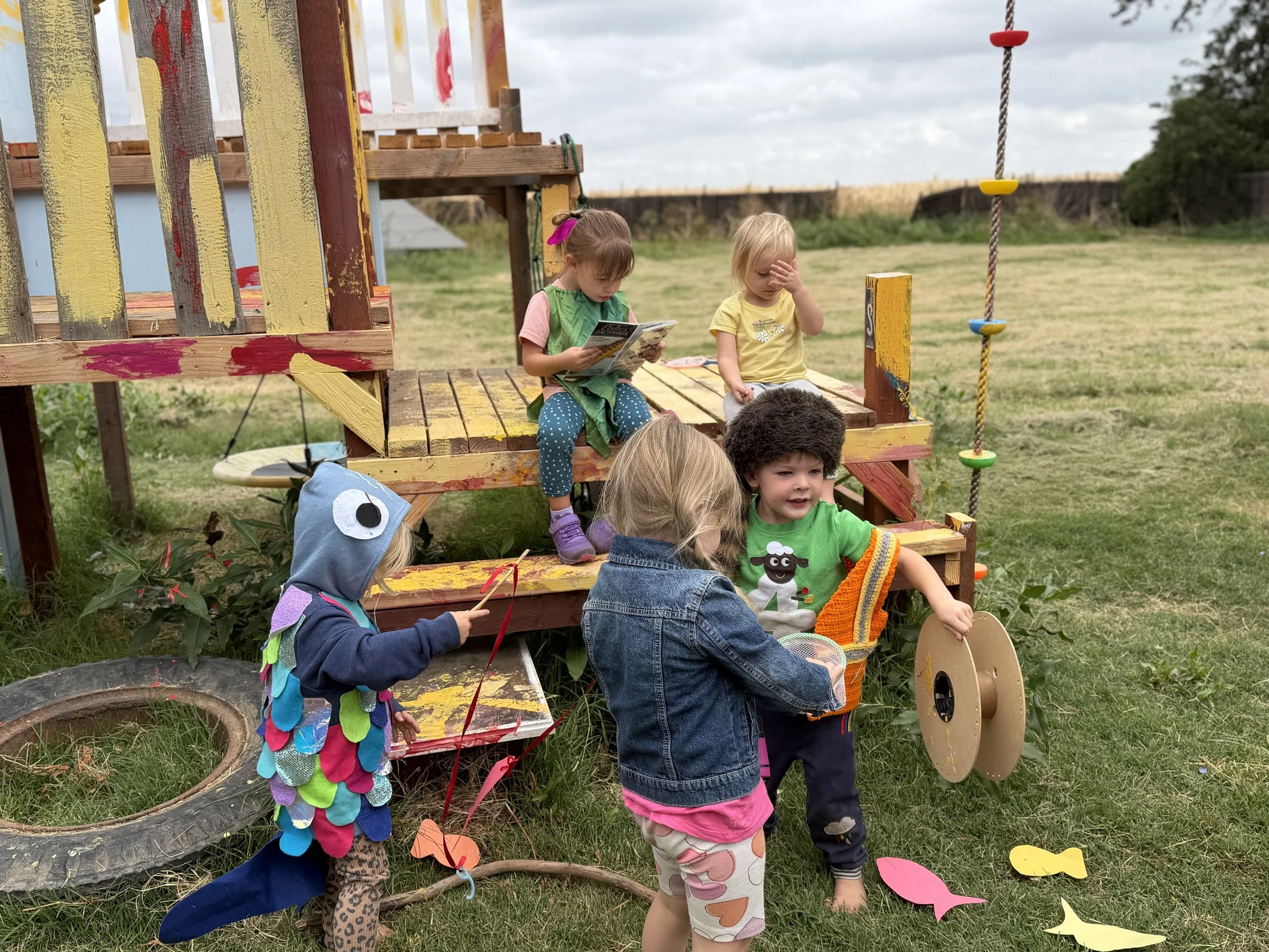 Kids playing on a colorful wooden playground structure outdoors, with some children dressed in costumes and others in casual clothing, with grassy field and cloudy sky in the background.
