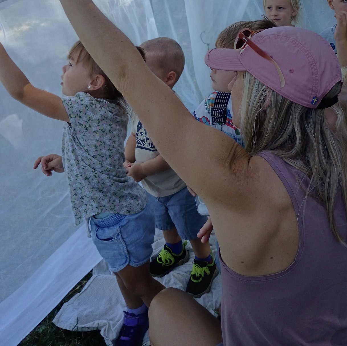 A woman and several children examining something inside a semi-transparent plastic tunnel outdoors.