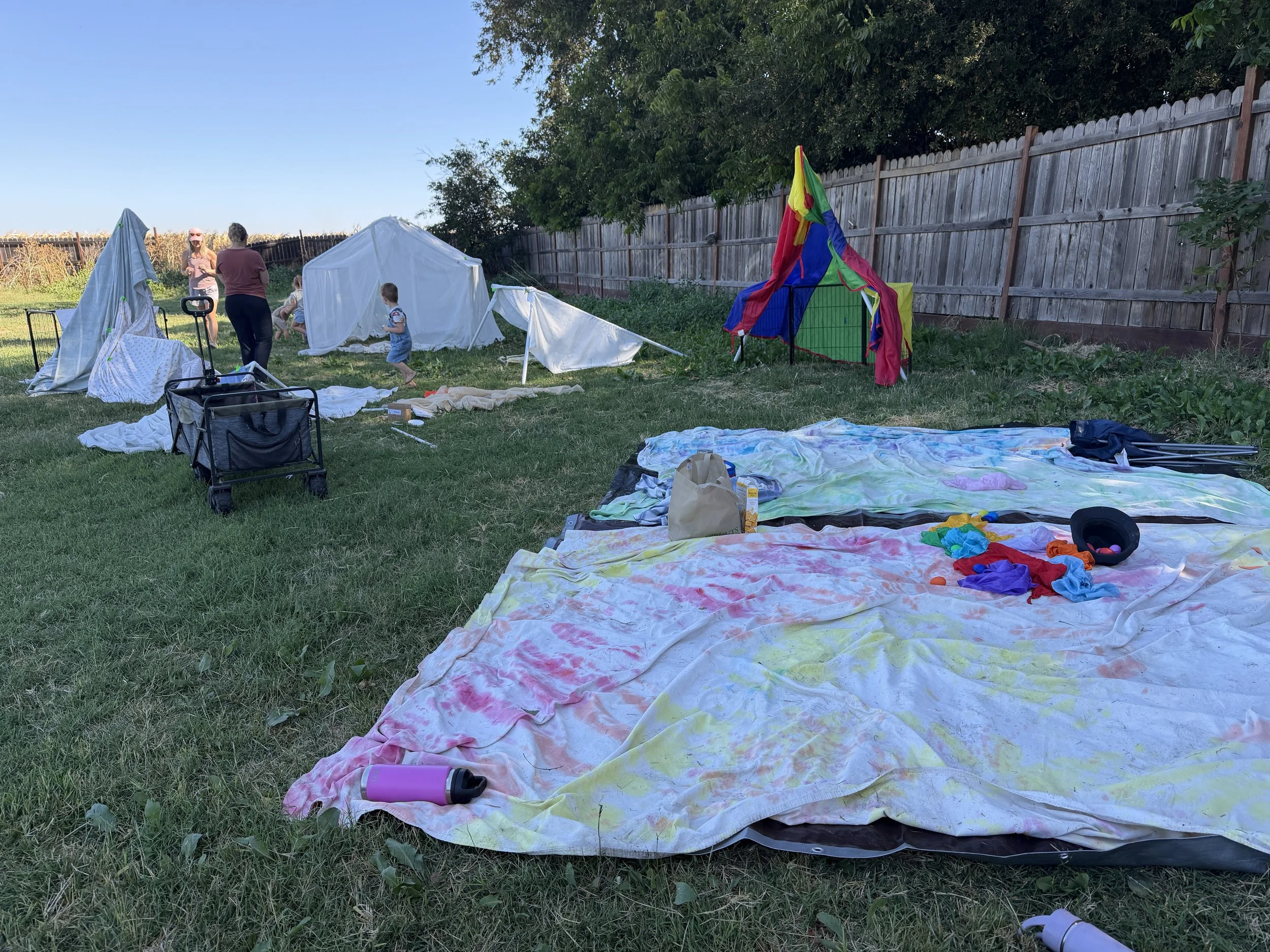 Backyard with grass, with colorful tie-dye blankets on the ground, a pink water bottle, and scattered toys. Two children and two adults are setting up a small white tent and an outdoor play area next to a wooden fence, with trees in the background an