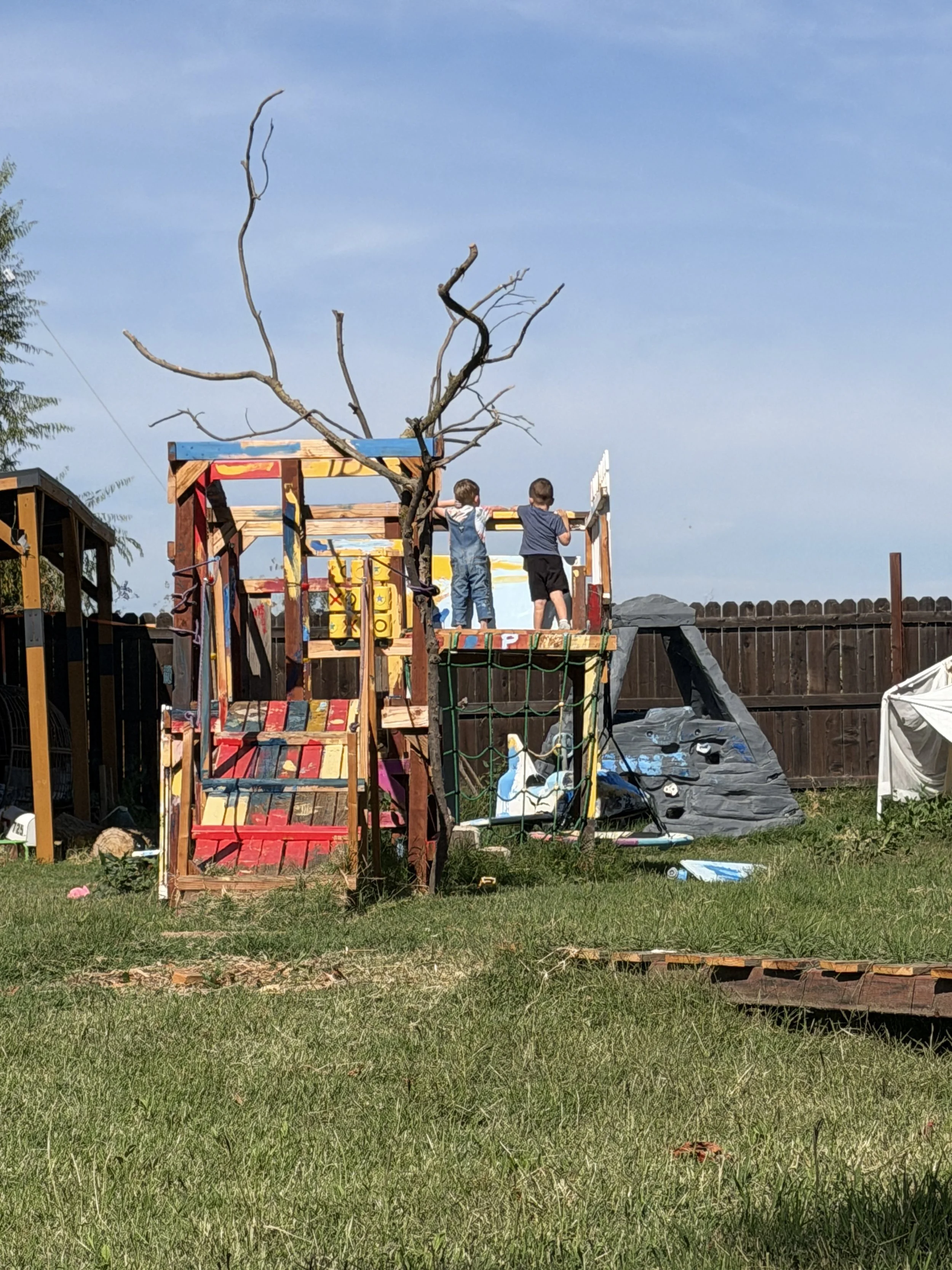 Two children standing on a colorful, wooden playhouse in a backyard, with a leafless tree and a wooden fence in the background.