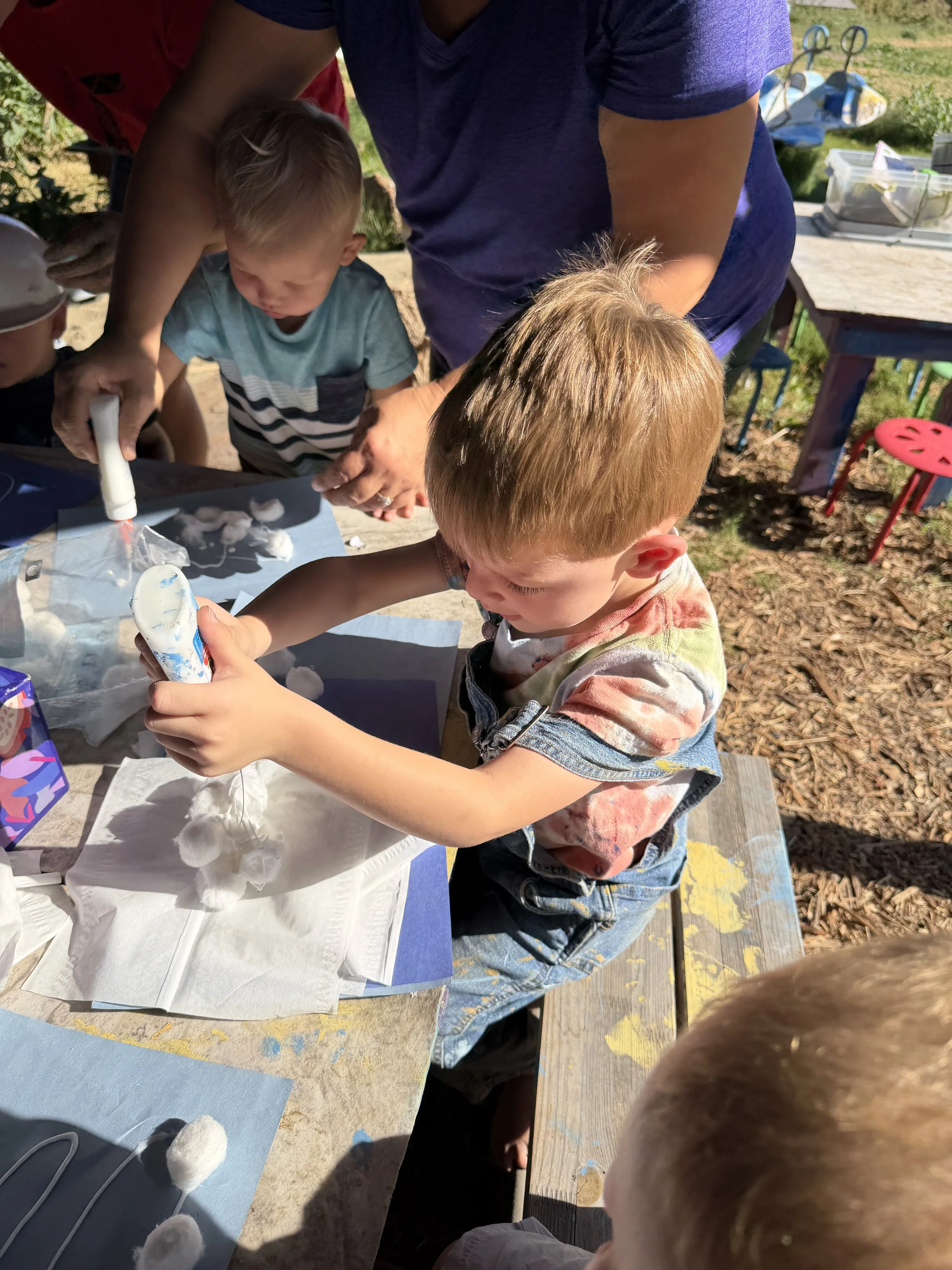 Children and adult engaging in outdoor activity, making cotton candy at a table with supplies, in a park or backyard setting.