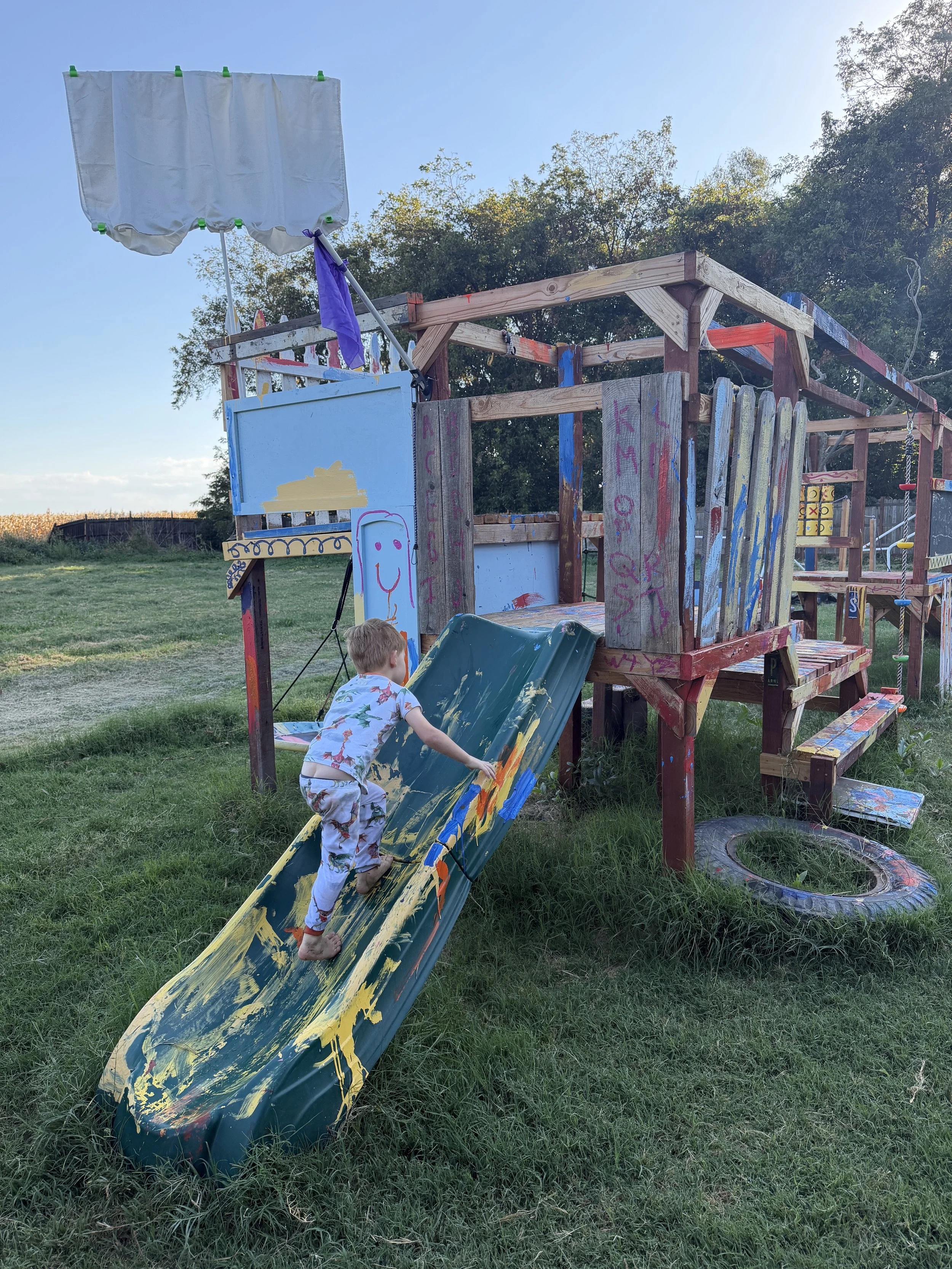 Child in pajamas playing on a painted outdoor slide at a backyard playground, with a wooden jungle gym and tire swing, under a clear blue sky.