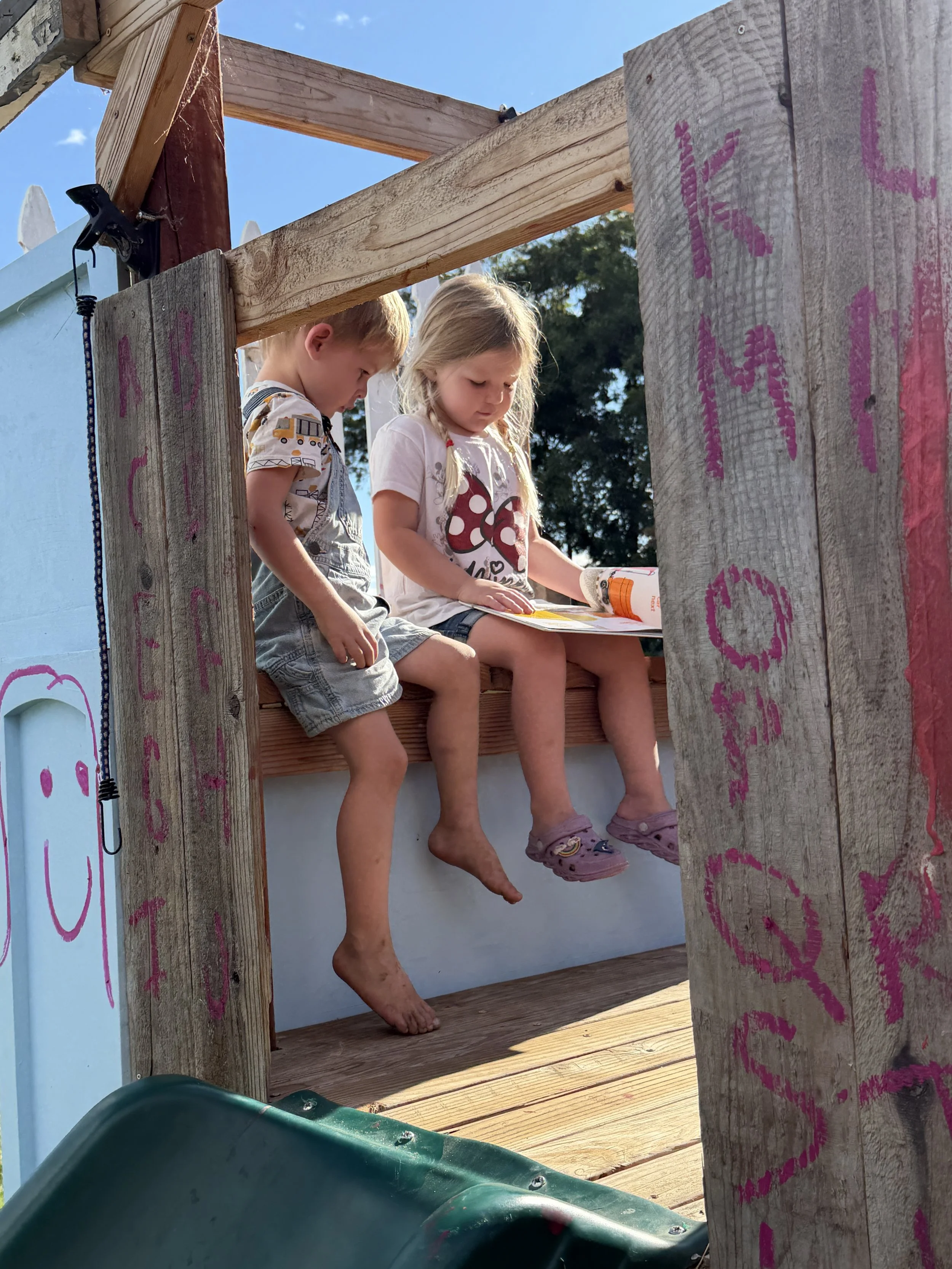 Two children sitting at the top of a wooden play structure with a slide, reading a book in an outdoor setting with blue sky and trees in the background.