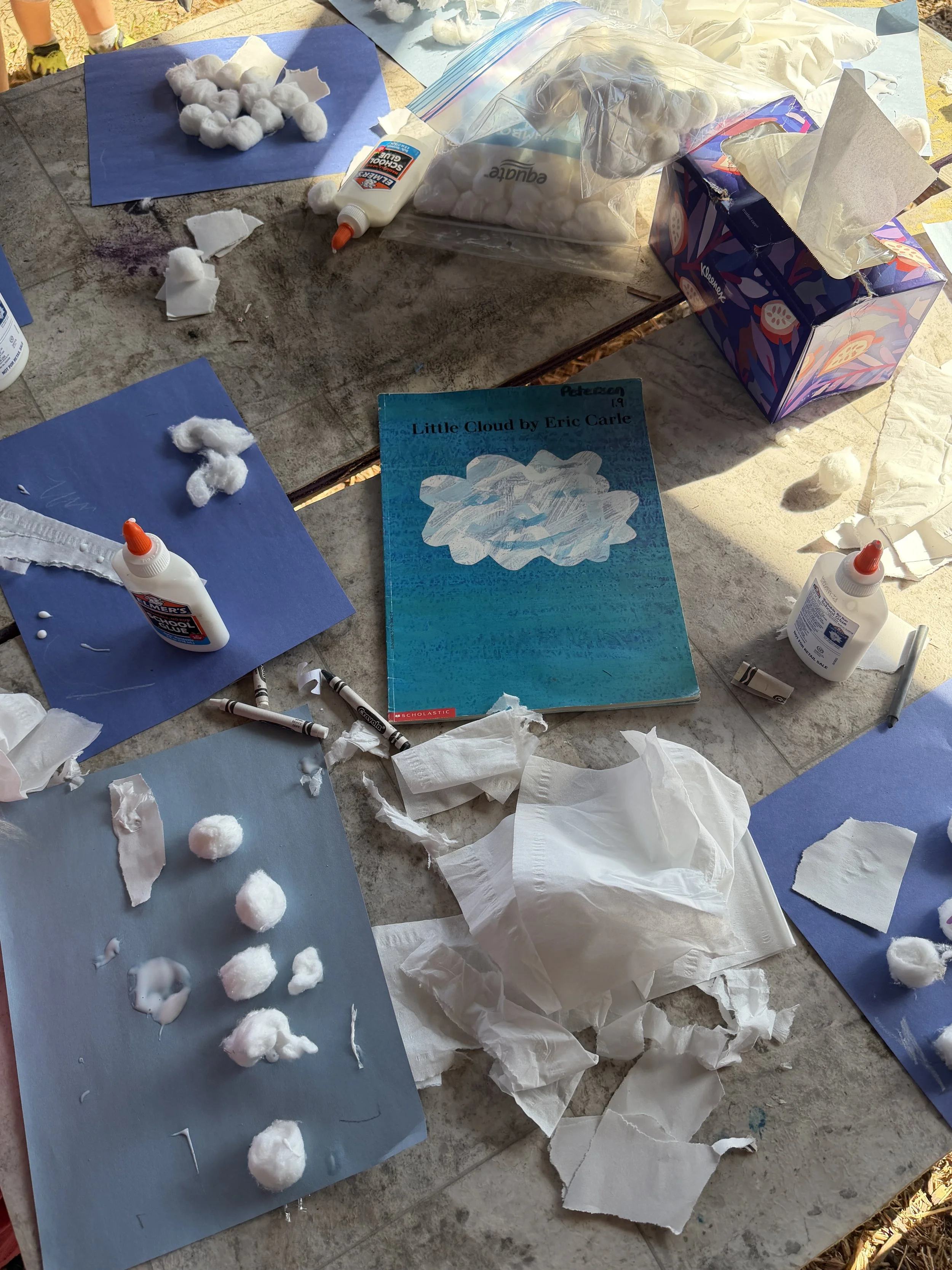 A table with art supplies for creating a cotton cloud craft, including cotton balls, glue, paper, and a craft book titled 'Little Cloud' by Eric Carle.
