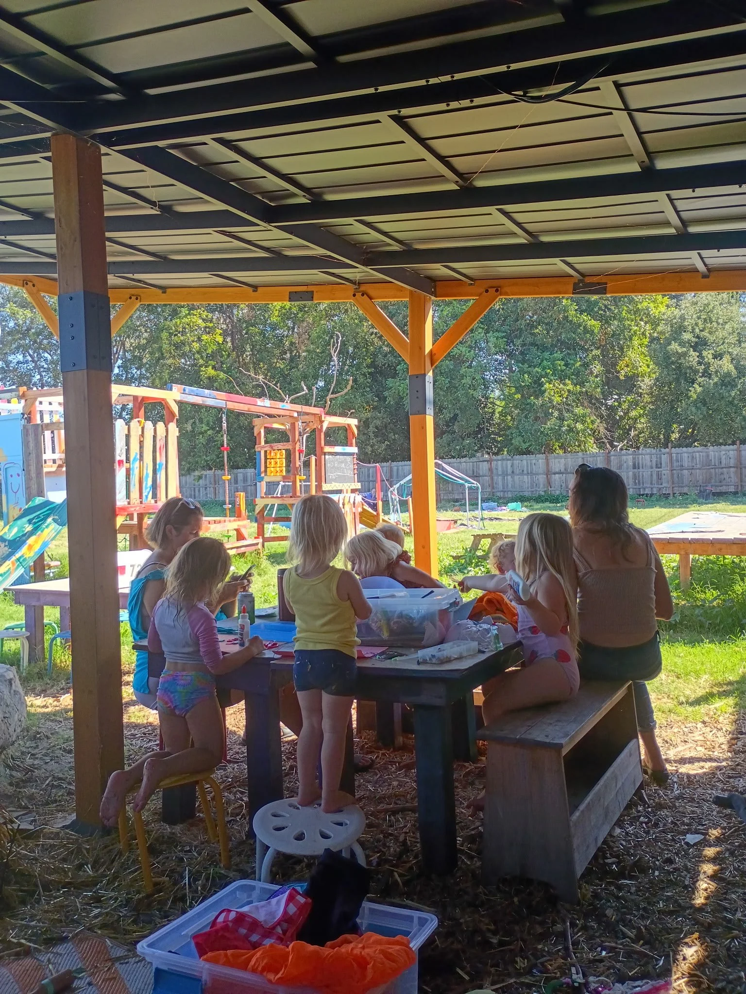 Children and adults sitting outside under a covered shelter, engaging in arts and crafts. There is playground equipment and a wooden fence in the background, with green trees and clear sky visible.