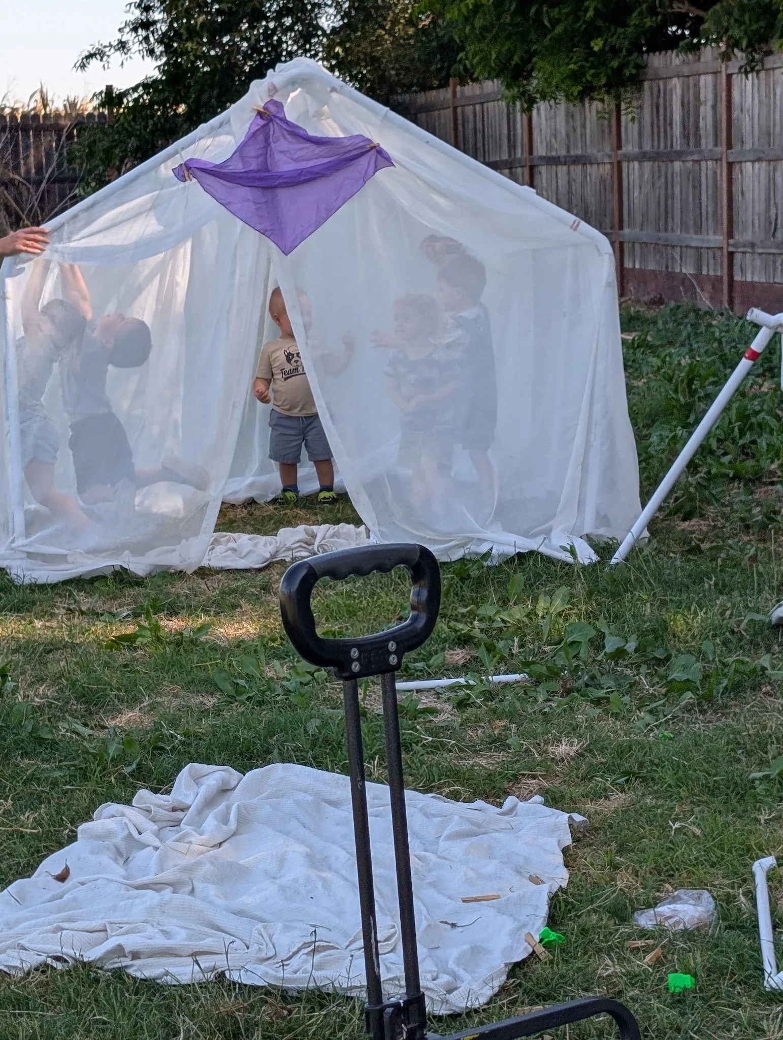 A butterfly-shaped fabric with purple wings appears to be a kite, set up outdoors in a backyard with children playing inside a white cloth enclosure, and a fence and trees in the background.