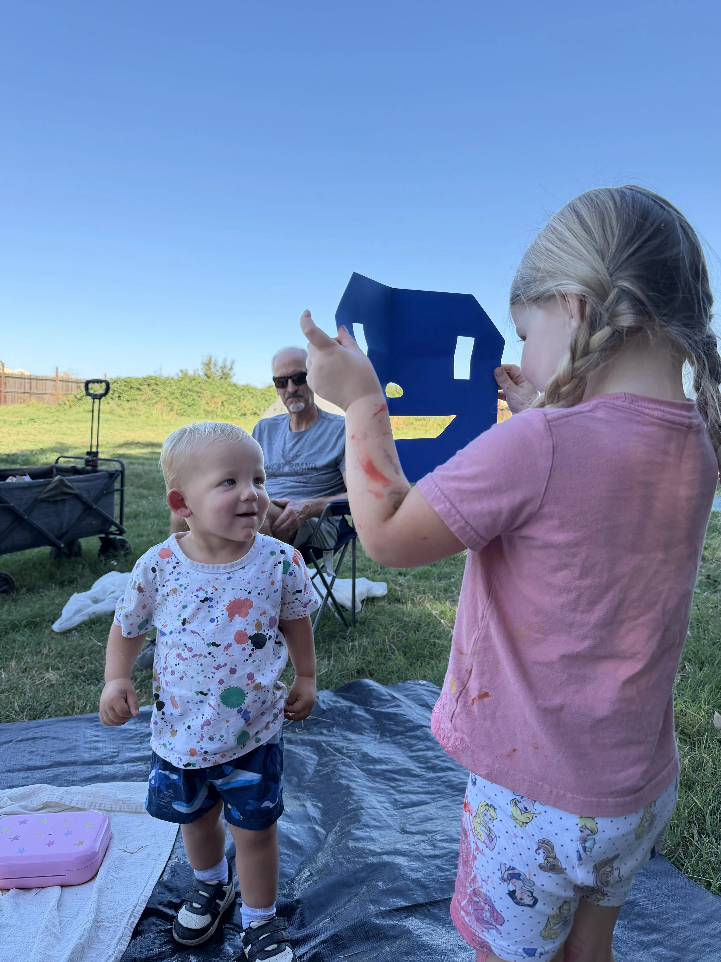 A young girl with blonde hair in braids and a pink shirt is holding a dark blue cutout of a whale, showing it to a young boy with blonde hair, a white shirt with colorful splatters, and shorts with a cartoon character pattern. An older man with sungl