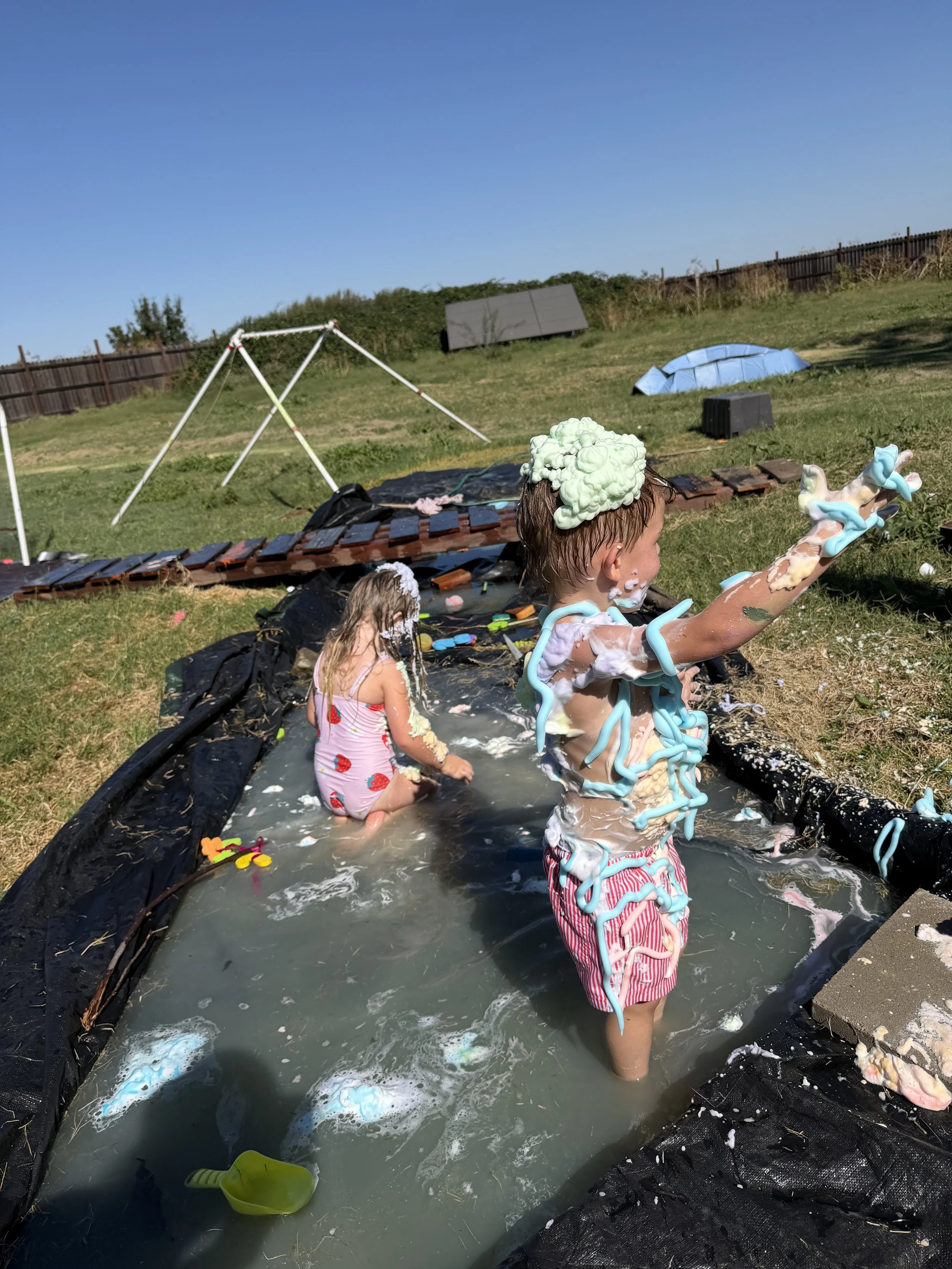 Children playing and covered in foam while standing in a small inflatable pool outdoors on a sunny day.
