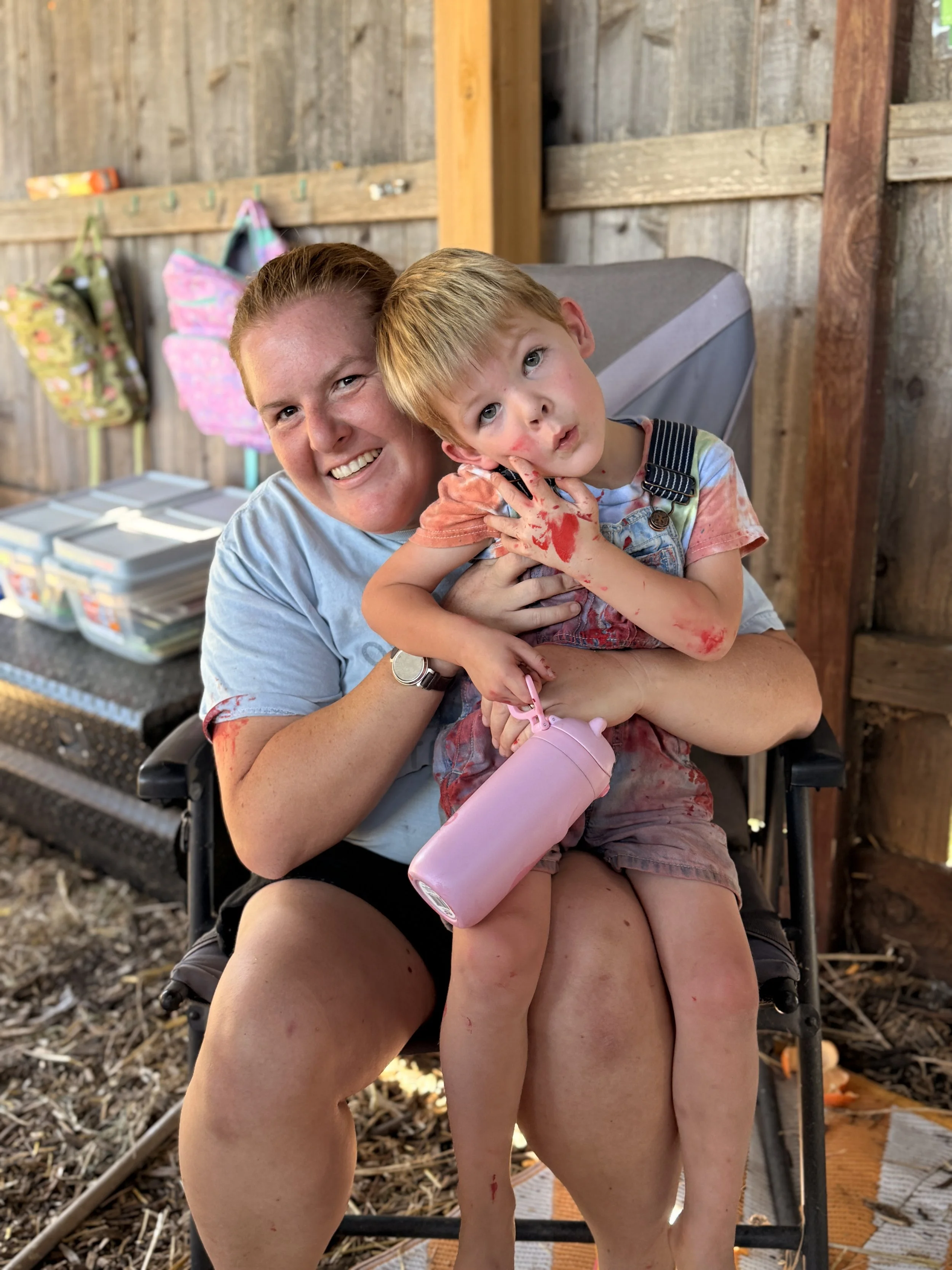 A woman and a young boy sitting on a chair, with the woman smiling and the boy looking at the camera with a neutral expression, both covered in red and pink paint or ink, in a rustic setting with wood walls and hanging bags in the background.