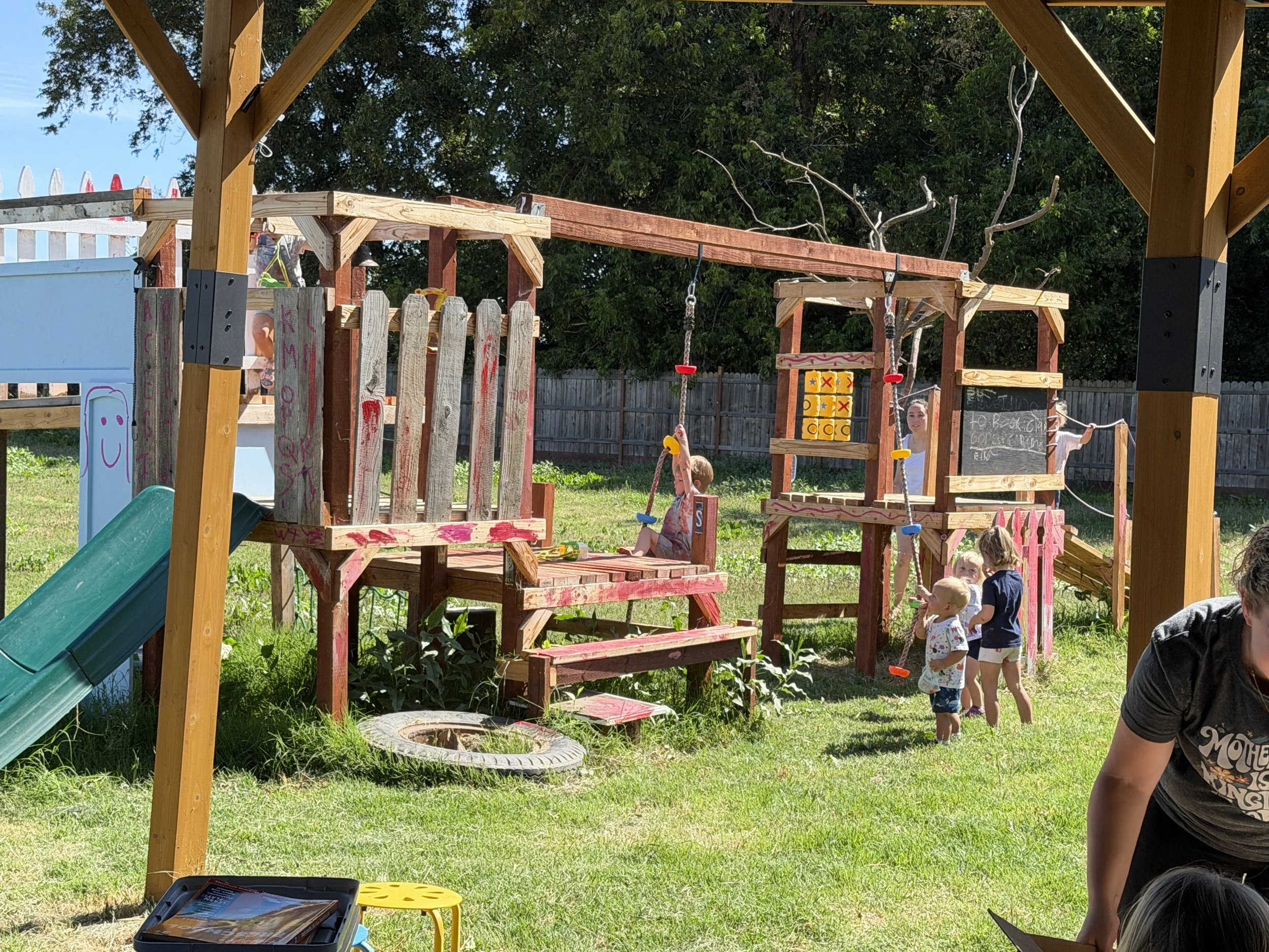 A children's playground made of wood with a slide, climbing structures, and swings. Several children and adults are present, with children playing and some standing in a line. The background shows a wooden fence and green trees.
