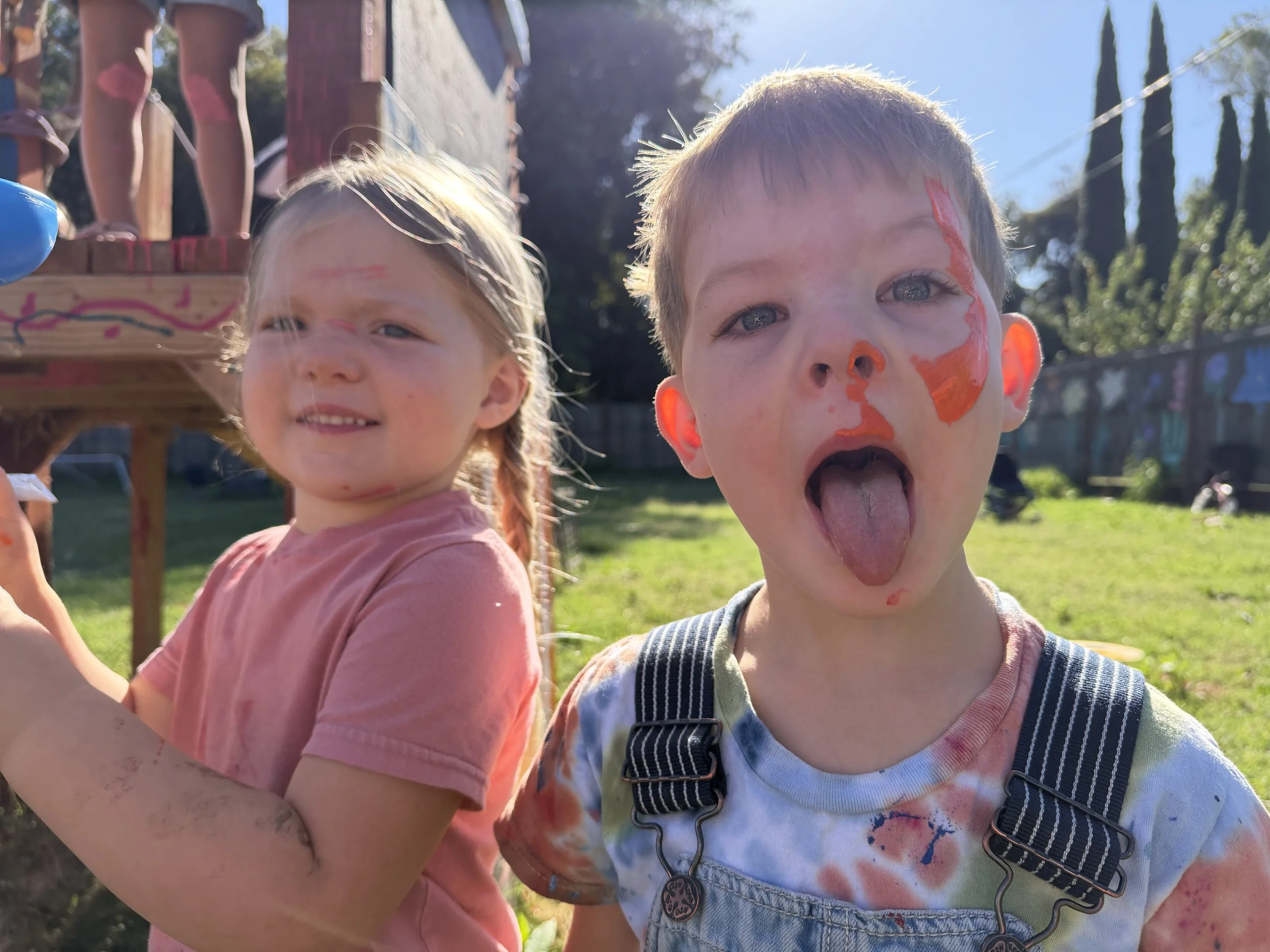 Two children with paint on their faces and clothes, standing outdoors near a wooden play structure with a rope ladder.