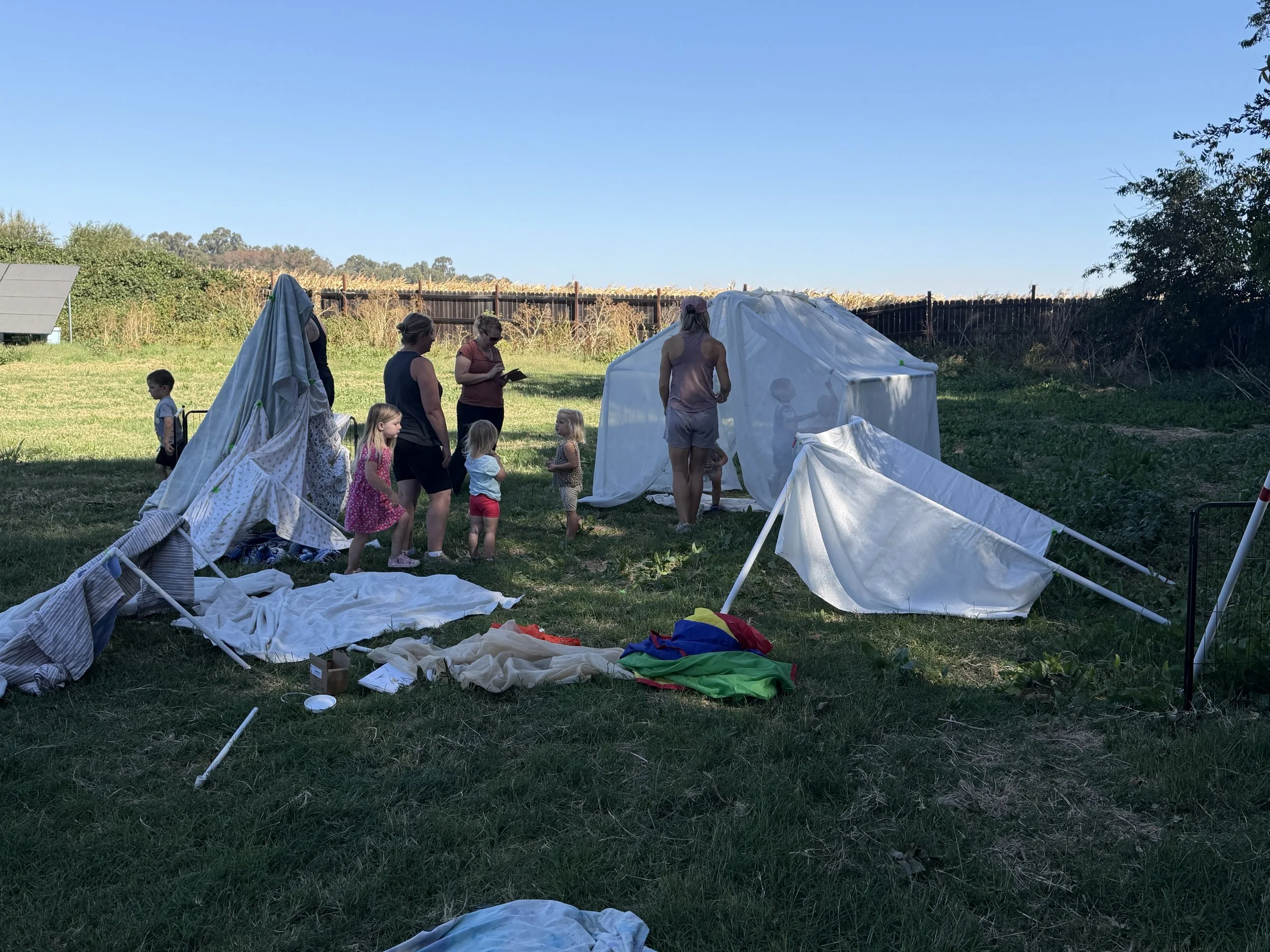 A group of children and adults standing outdoors on a grassy area next to a white tent structure, with supplies and fabric scattered around. There is a clear blue sky and a wooden fence in the background.