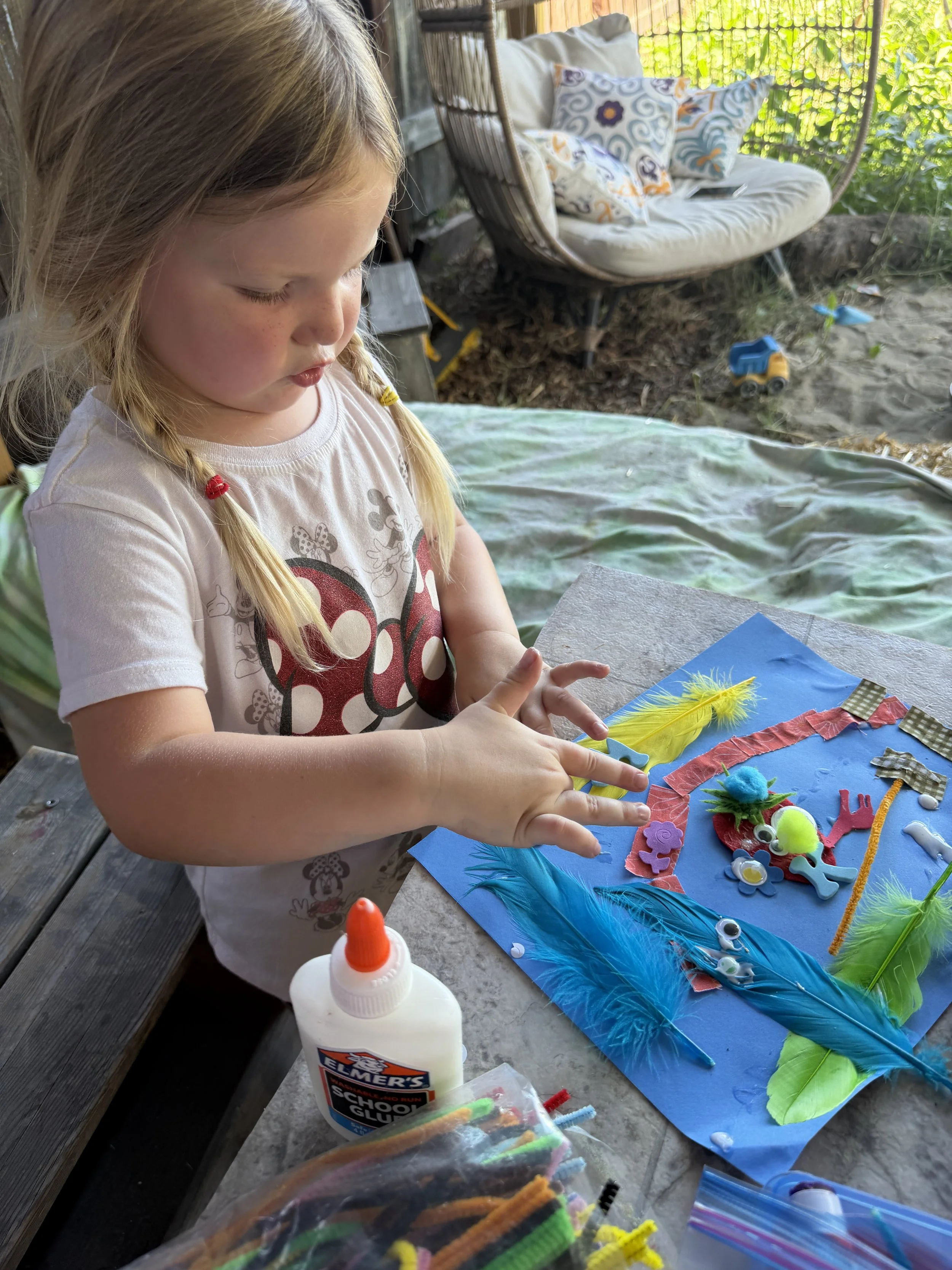 A young girl with blonde hair in braids is sitting at a table, working on a craft project with colorful feathers, foam shapes, and glue. There are craft supplies like glue bottles and pipe cleaners on the table, and a wooden porch with a hanging chai