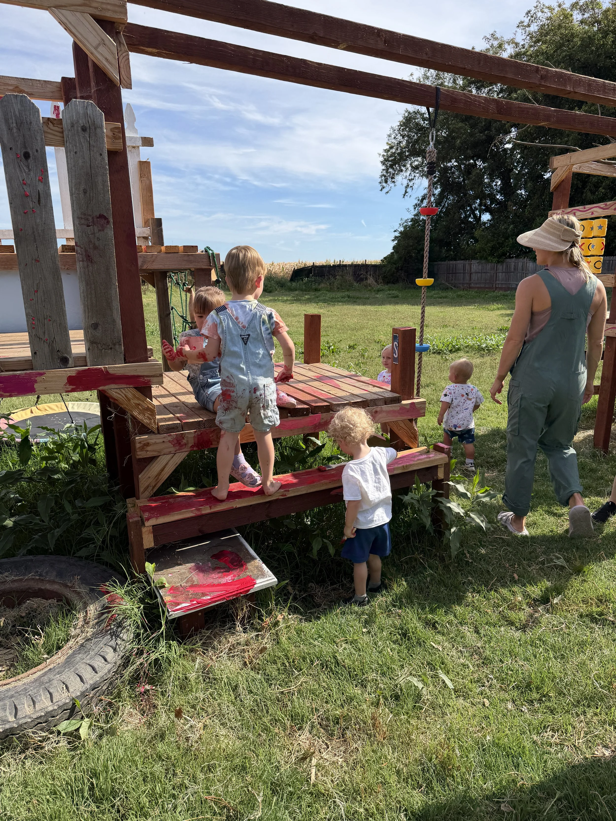 Children playing on a wooden play structure with adults supervising in a grassy backyard on a sunny day.