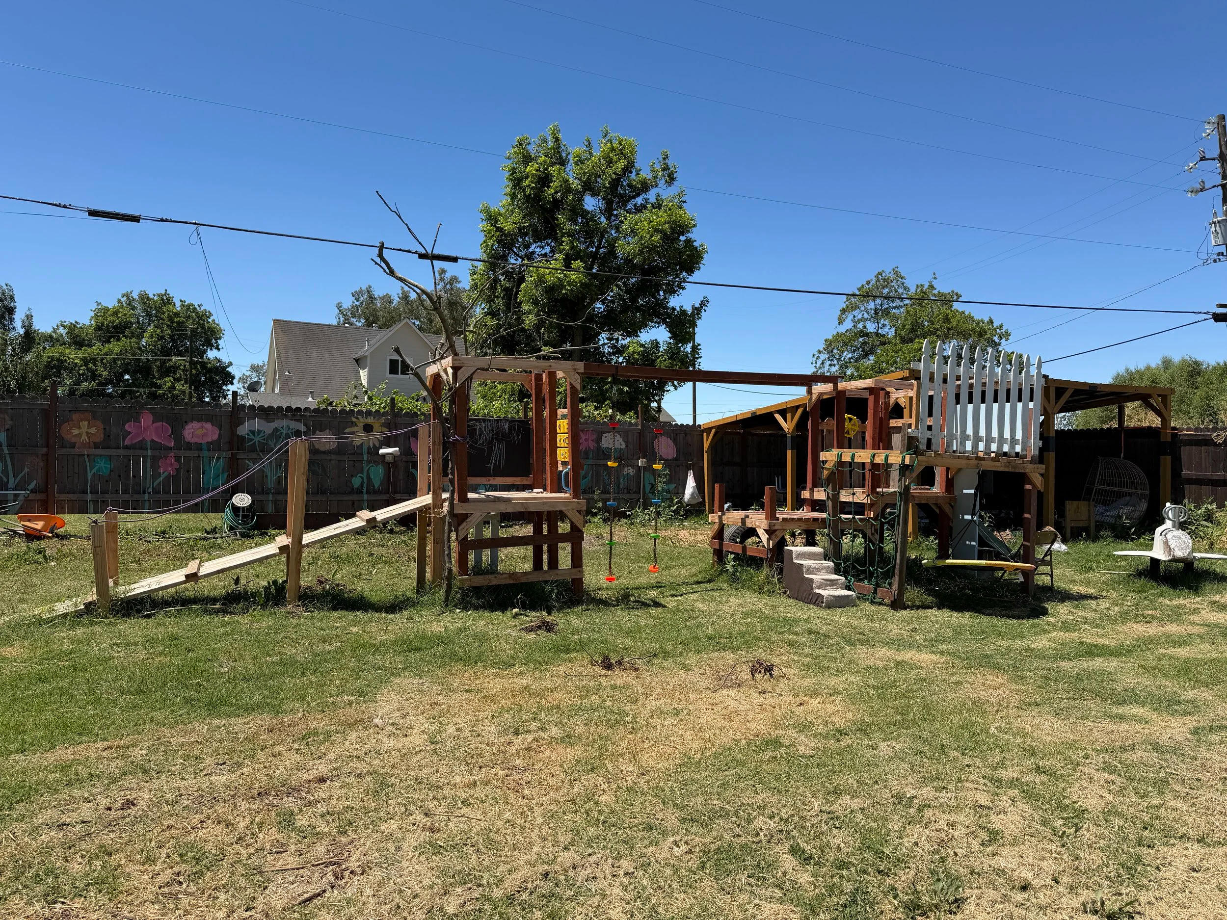 Backyard with a wooden play structure, a white picket fence, a tree, and a mural on the fence featuring flowers. Clear blue sky and rooftops of neighboring houses visible in the background.