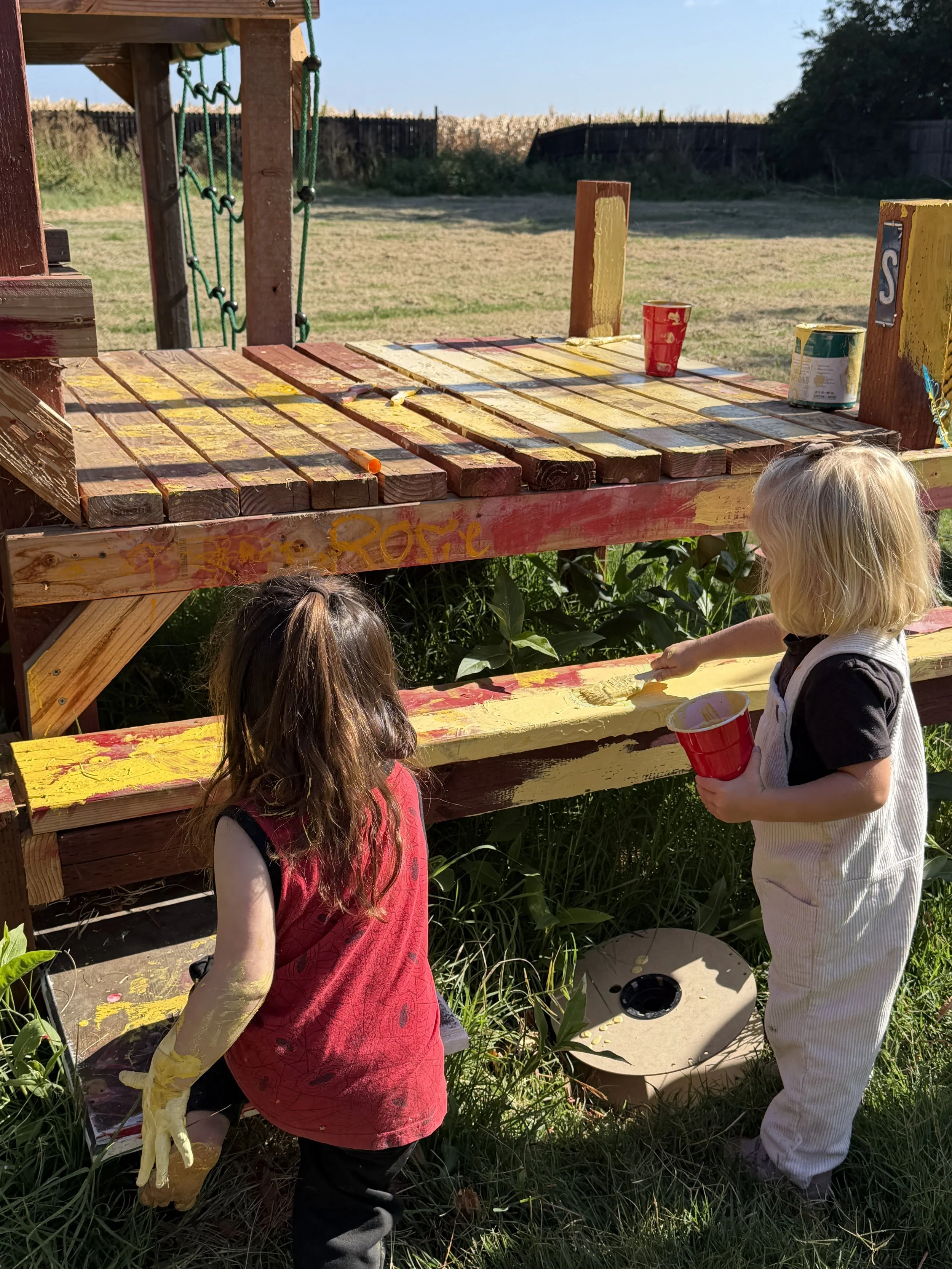 Three children painting a wooden structure outside on a sunny day. The structure is covered in yellow paint.
