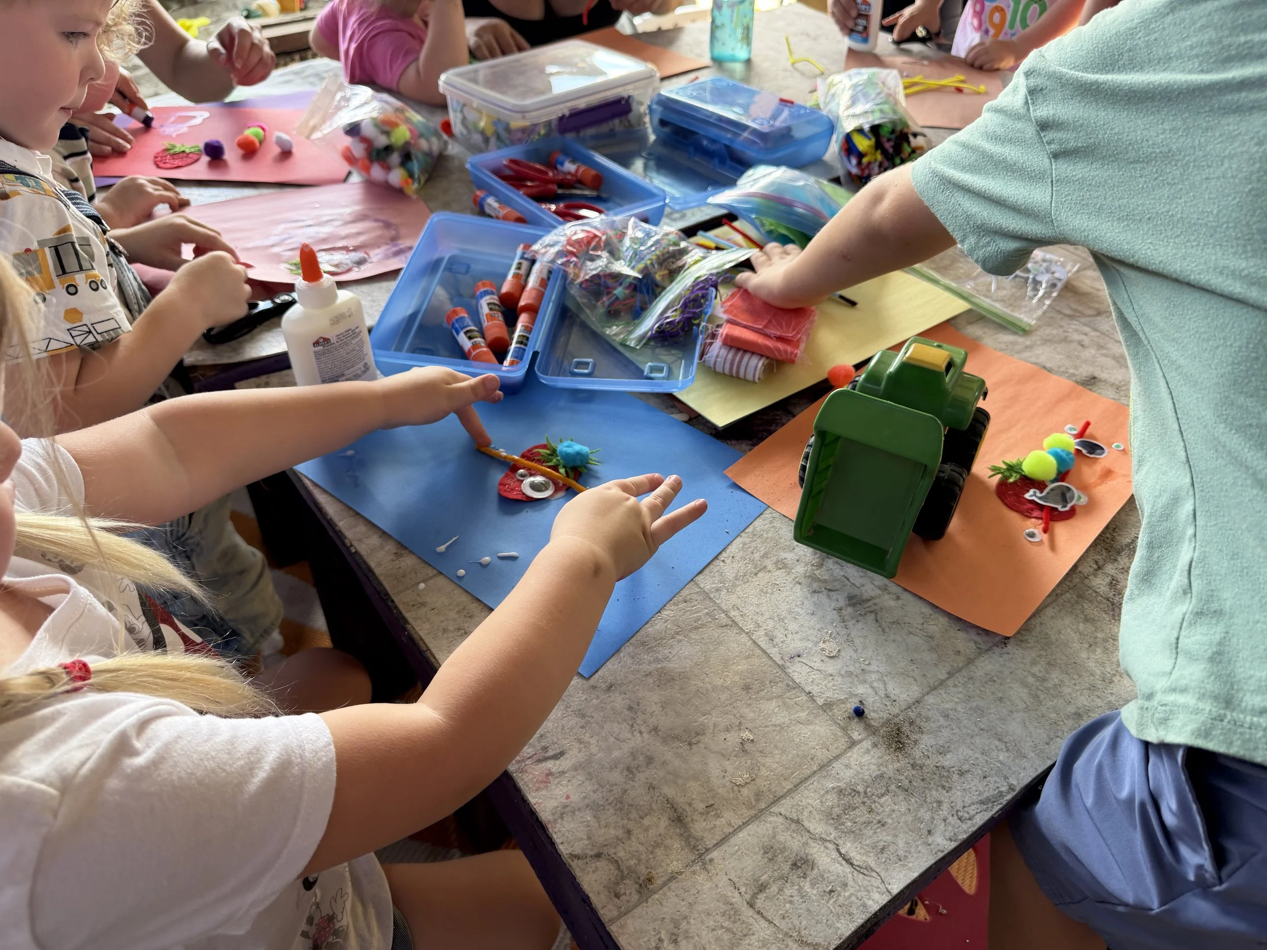 Children gathered around a table engaging in arts and crafts, with various craft supplies such as glue, scissors, markers, pom-poms, and paper on the table.