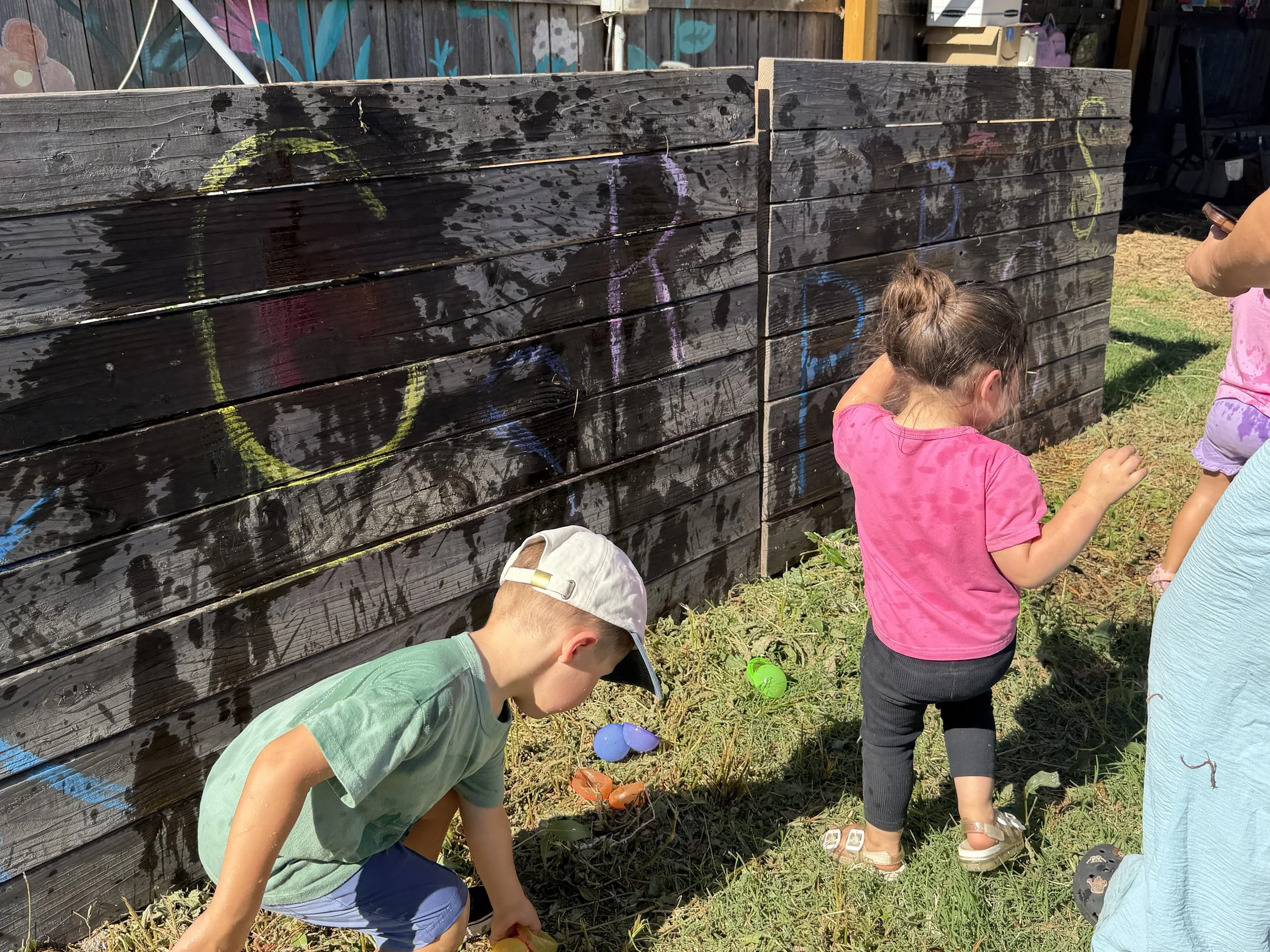 Children participating in an Easter egg hunt outside, gathering colorful plastic eggs near a painted wooden fence with graffiti.
