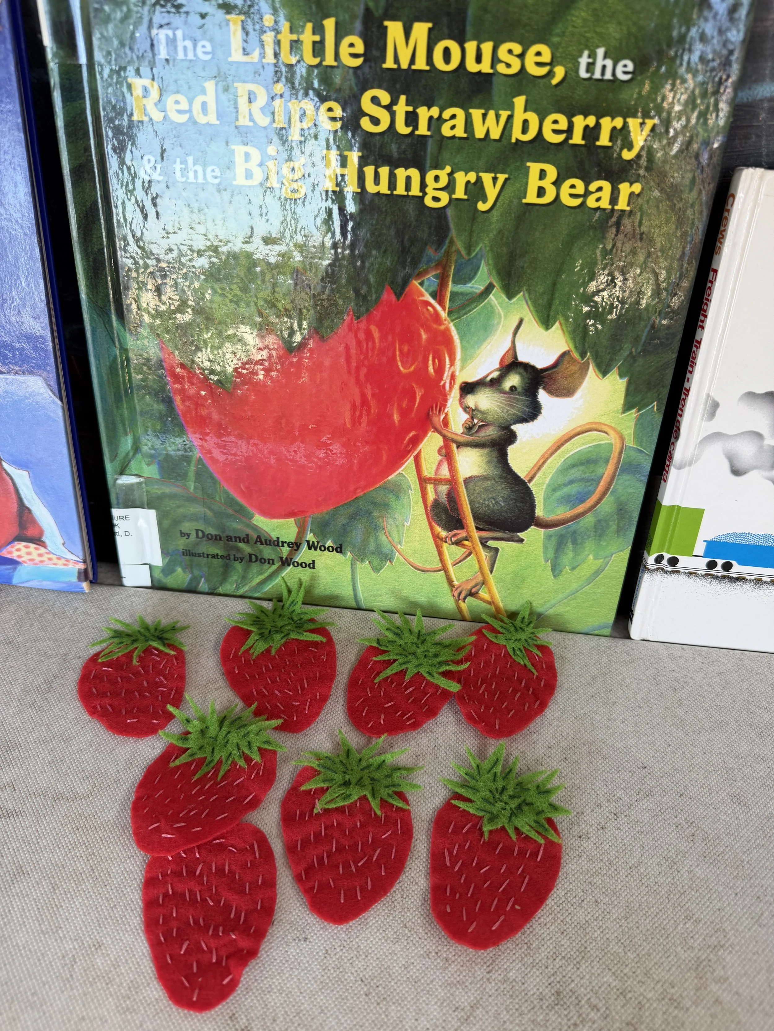A pile of red felt strawberries with green felt leaves and white stitching, placed in front of a children's book titled 'The Little Mouse, the Red Ripe Strawberry & the Big Hungry Bear' by Don and Audrey Wood. The strawberries are arranged on a beige