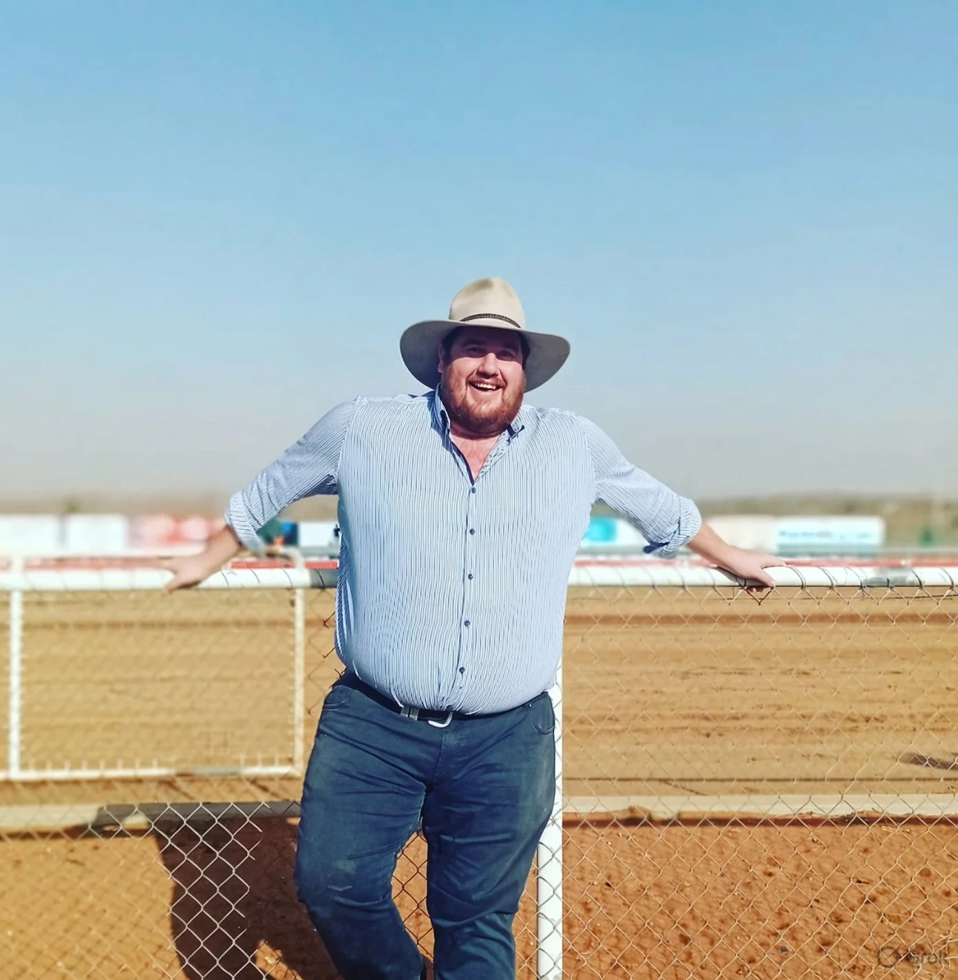 A man with a beard, wearing a wide-brimmed hat, a striped button-up shirt, and dark pants, stands smiling outdoors behind a chain-link fence, with a racetrack and open sky in the background.